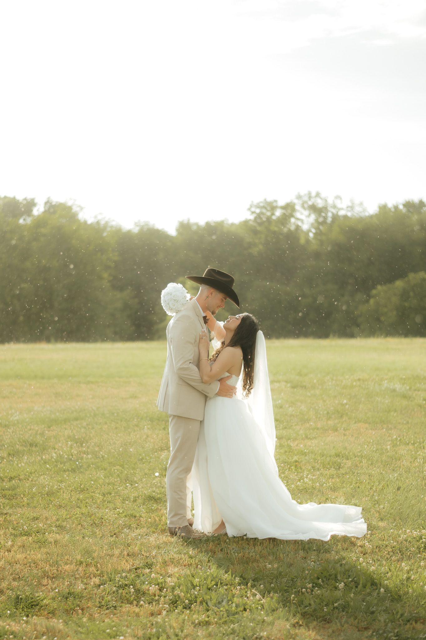 Bride and groom kissing in a sunlit field, groom wearing a cowboy hat and tan suit, bride in a flowing white gown.
