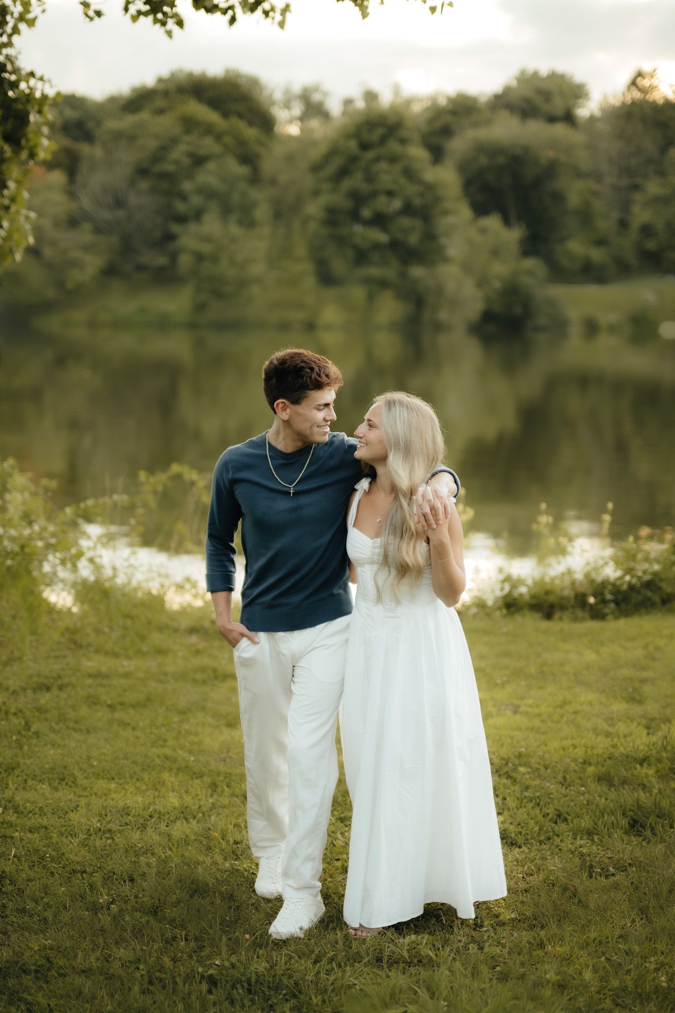 Young couple standing by a serene lake surrounded by lush green trees, woman in white dress and man in blue shirt.