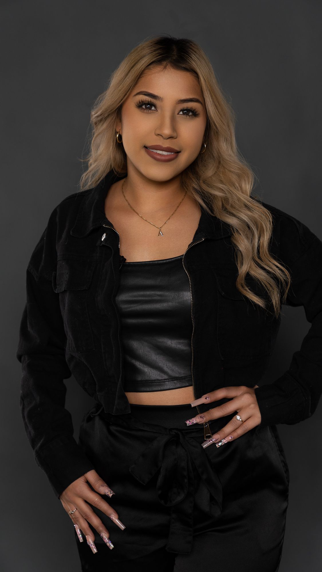 Professional portrait of someone wearing a black crop top, jacket and pants with long wavy blonde hair against dark background.