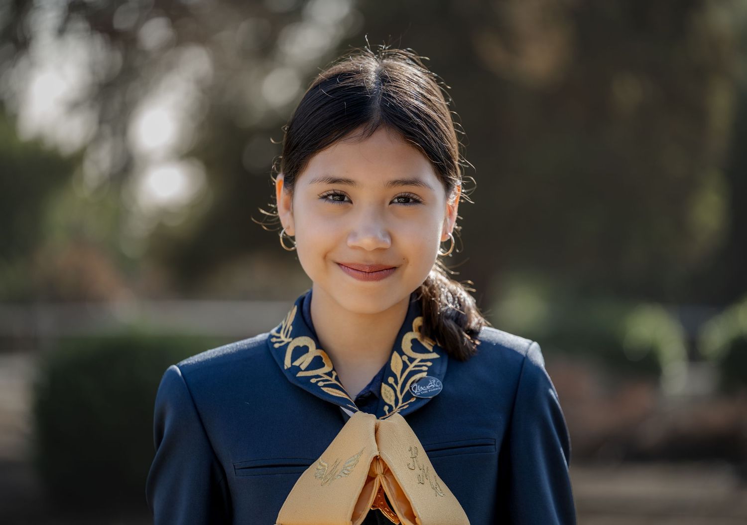 A person wearing a navy blue mariachi uniform with a patterned collar smiles at the camera in an outdoor setting.