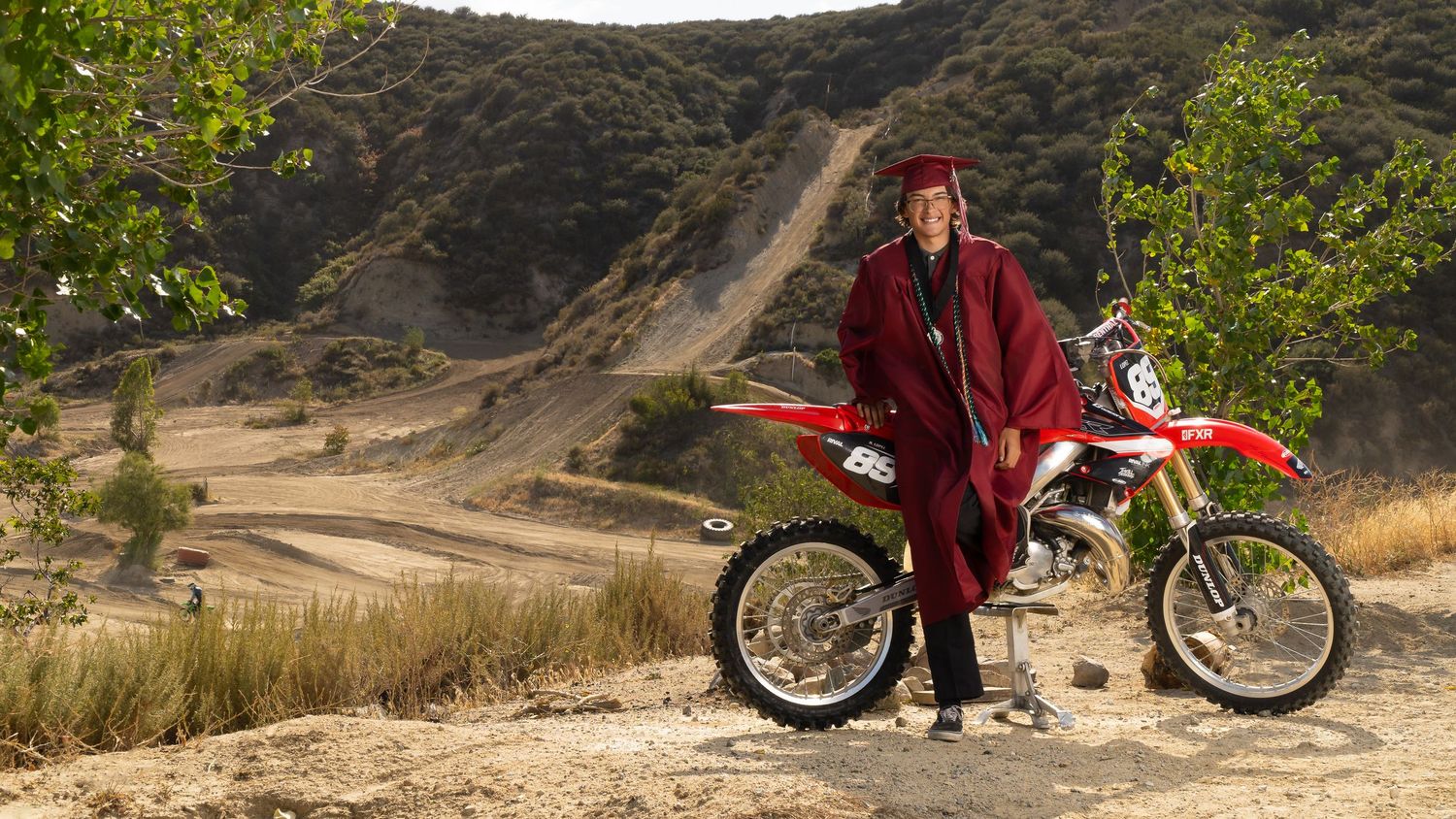 Graduate in cap and gown poses beside a red dirt bike on a sandy desert trail with hills in background.