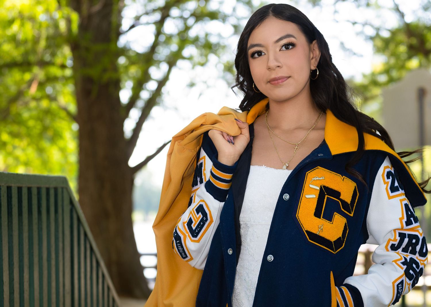 Young woman in navy and gold varsity letterman jacket posing outdoors for senior portrait photo.