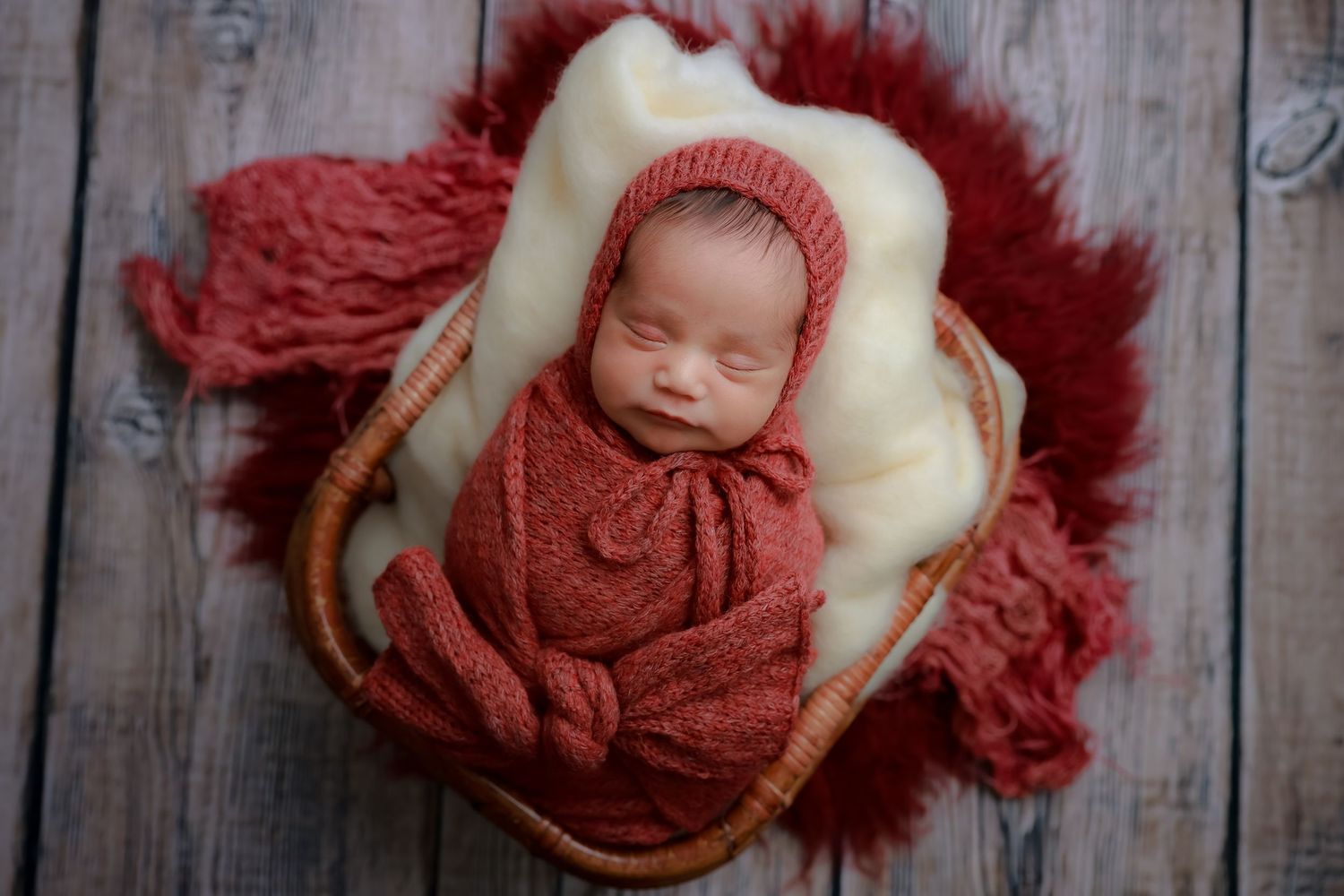 Sleeping newborn wrapped in rust-colored knit blanket nestled in cream basket with matching textured backdrop. luxury-family-photography