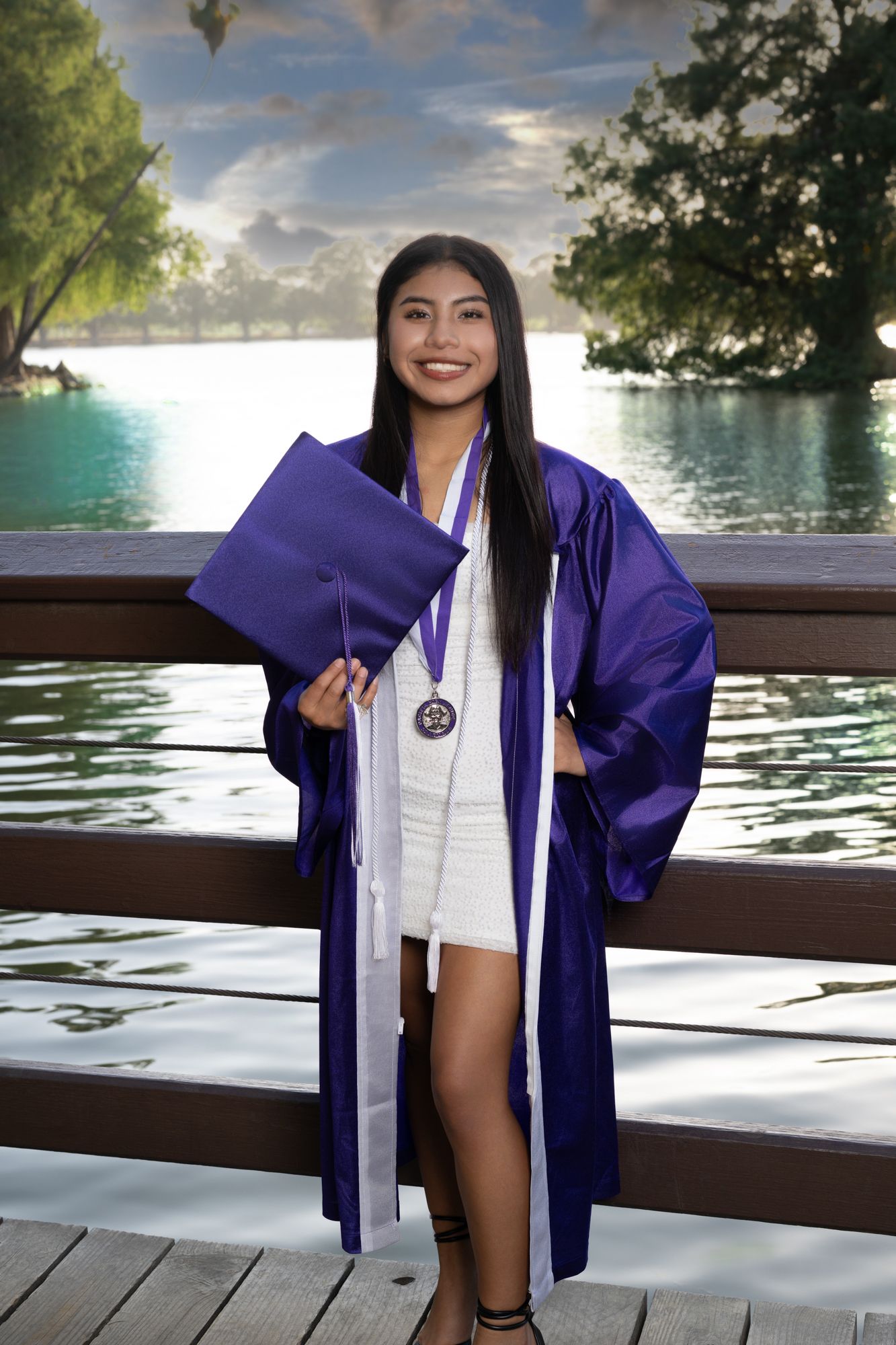 Graduate in purple and white academic regalia poses by a lake during a sunny graduation photoshoot.