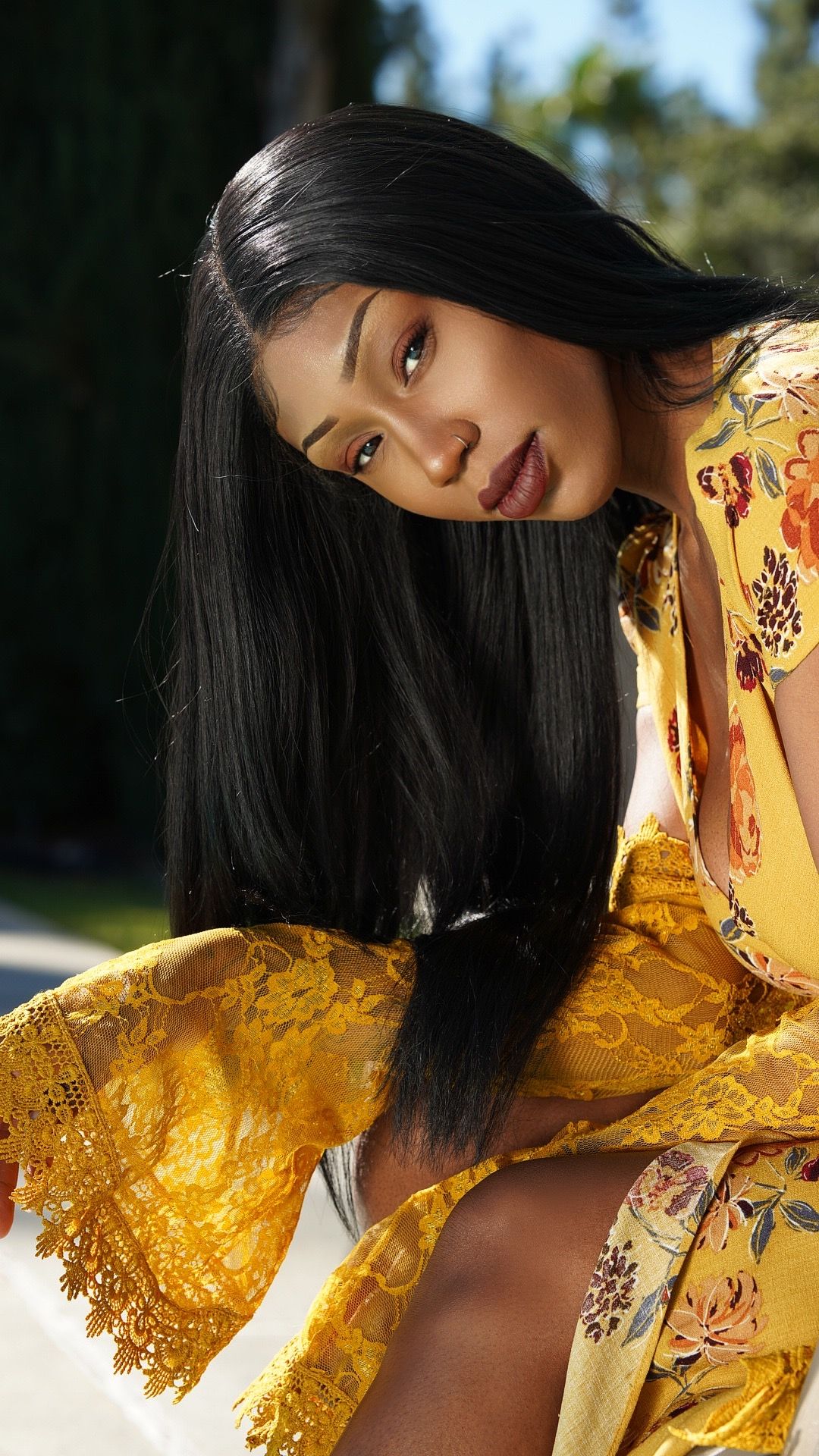 A person in a yellow floral dress with lace sleeves poses outdoors in natural sunlight.