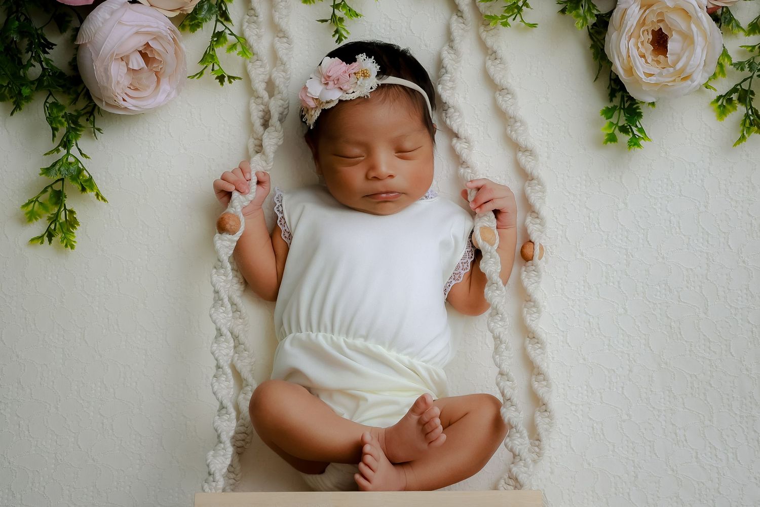 A sleepingvbaby in white clothing lies on a floral backdrop with pink roses and greenery. Corona luxury newborn photography experiene