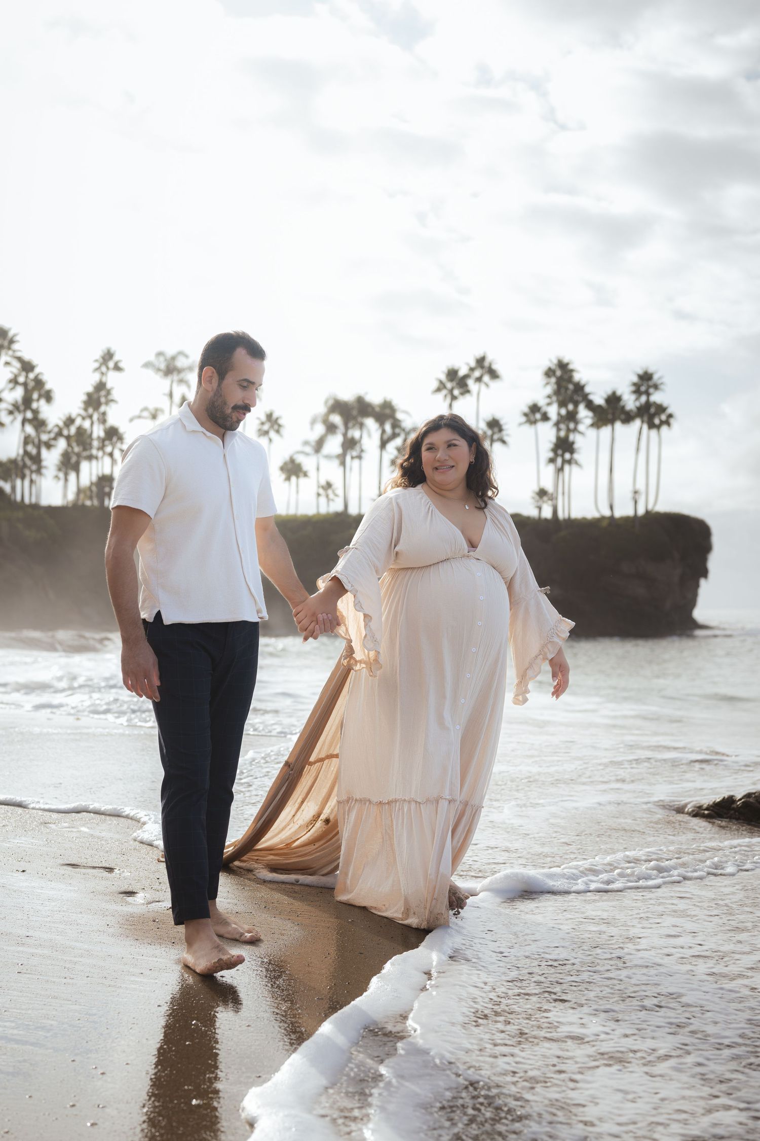 A couple walks hand in hand along a beach with palm trees and cliffs at sunset, waves gently lapping at their feet.