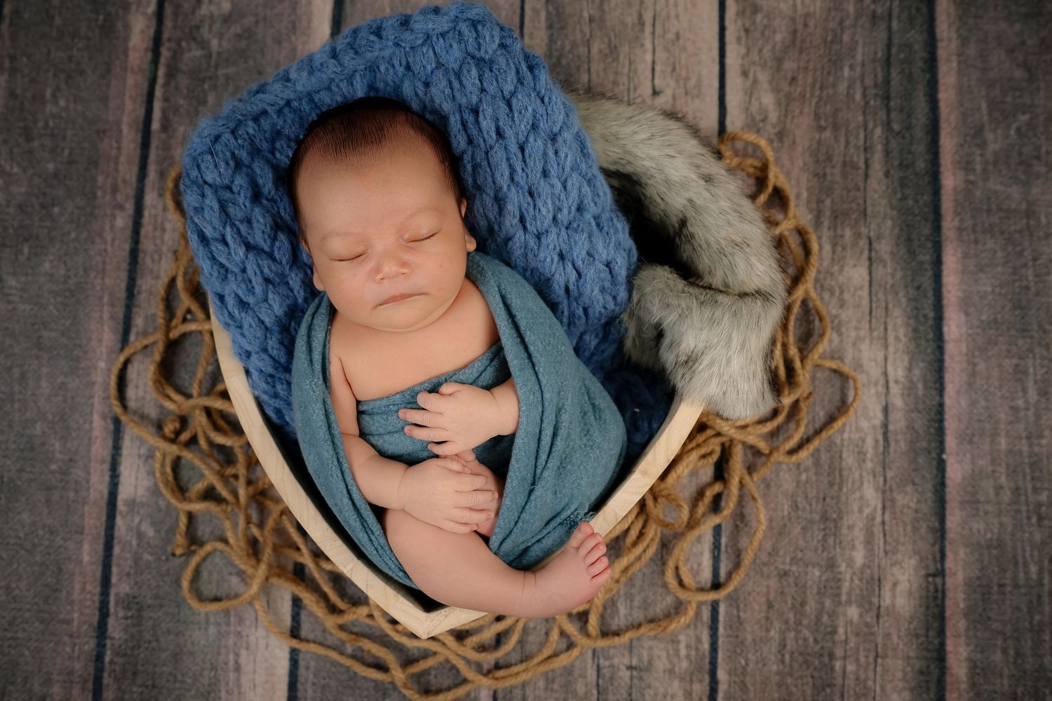 Sleeping newborn in blue wrap nestled in round woven basket against rustic wooden backdrop.