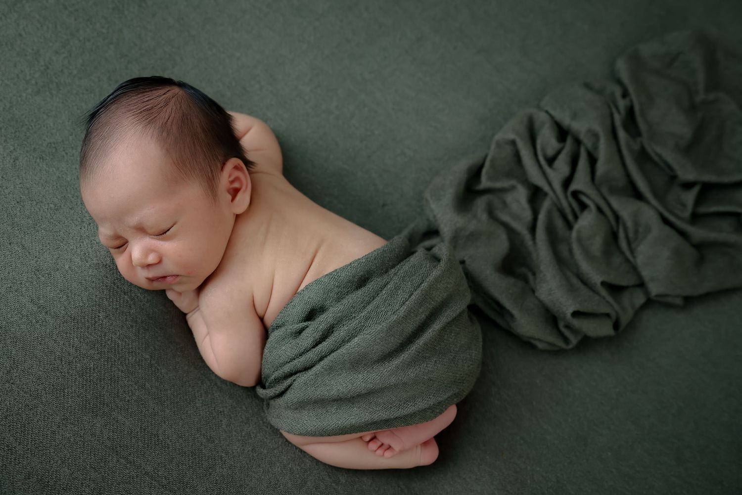 Sleeping newborn wrapped in olive green fabric against dark green textured backdrop.