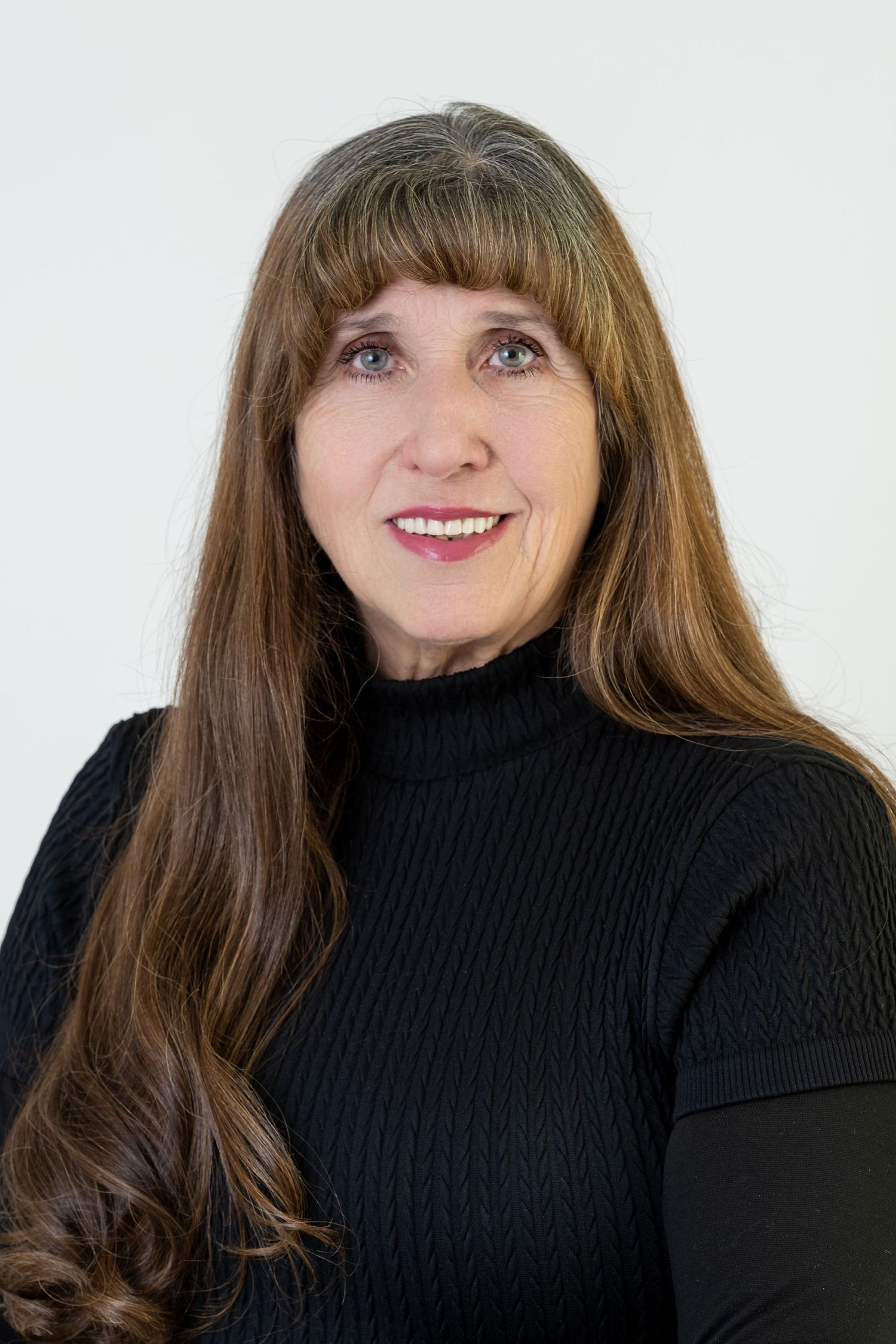 A person with long brown hair and bangs wearing a black turtleneck sweater poses against a white background.