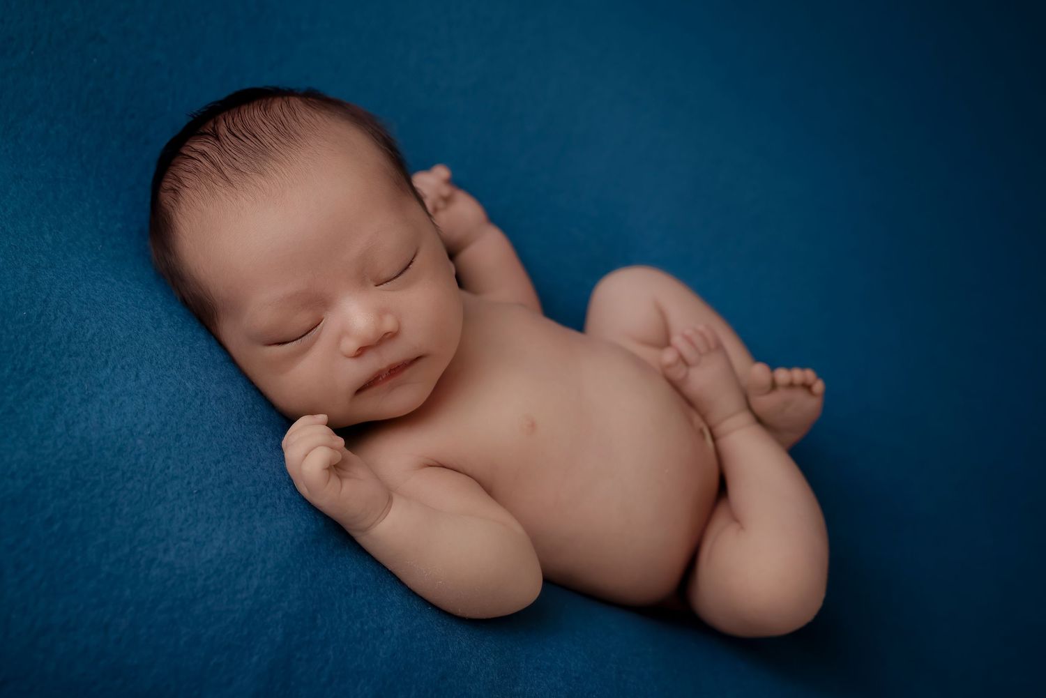 A sleeping newborn baby curled up on a dark blue textured background in a professional studio setting. luxury-family-photography-La-Puente