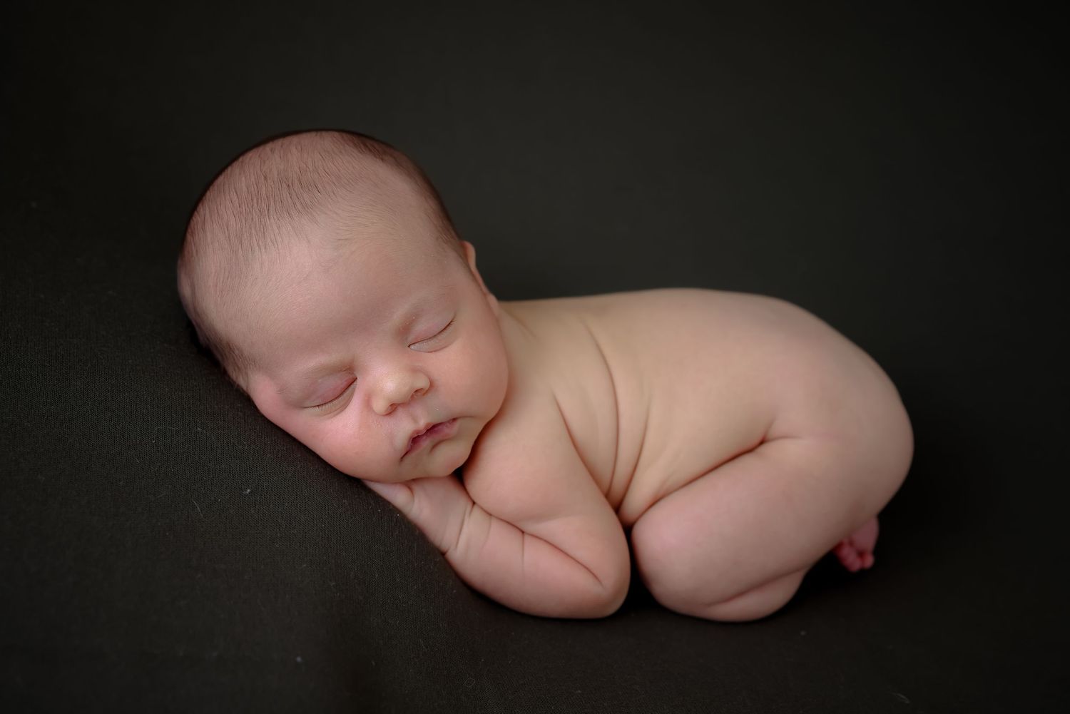 Peaceful sleeping newborn baby curled up on dark background in professional studio portrait.