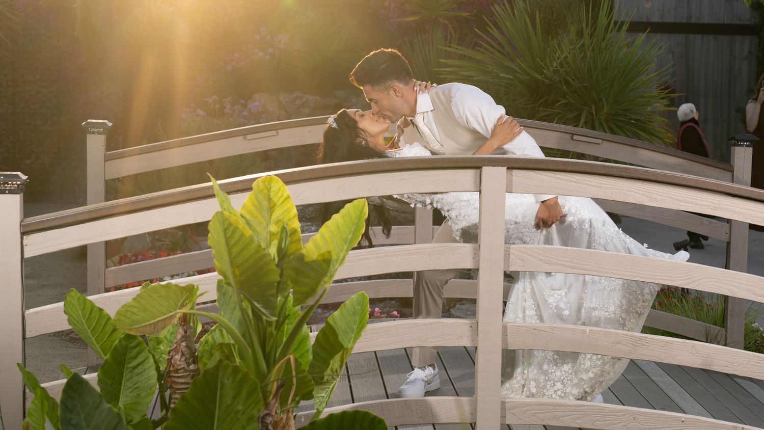 A bride and groom kiss on a white garden bridge at sunset with green plants in the foreground.