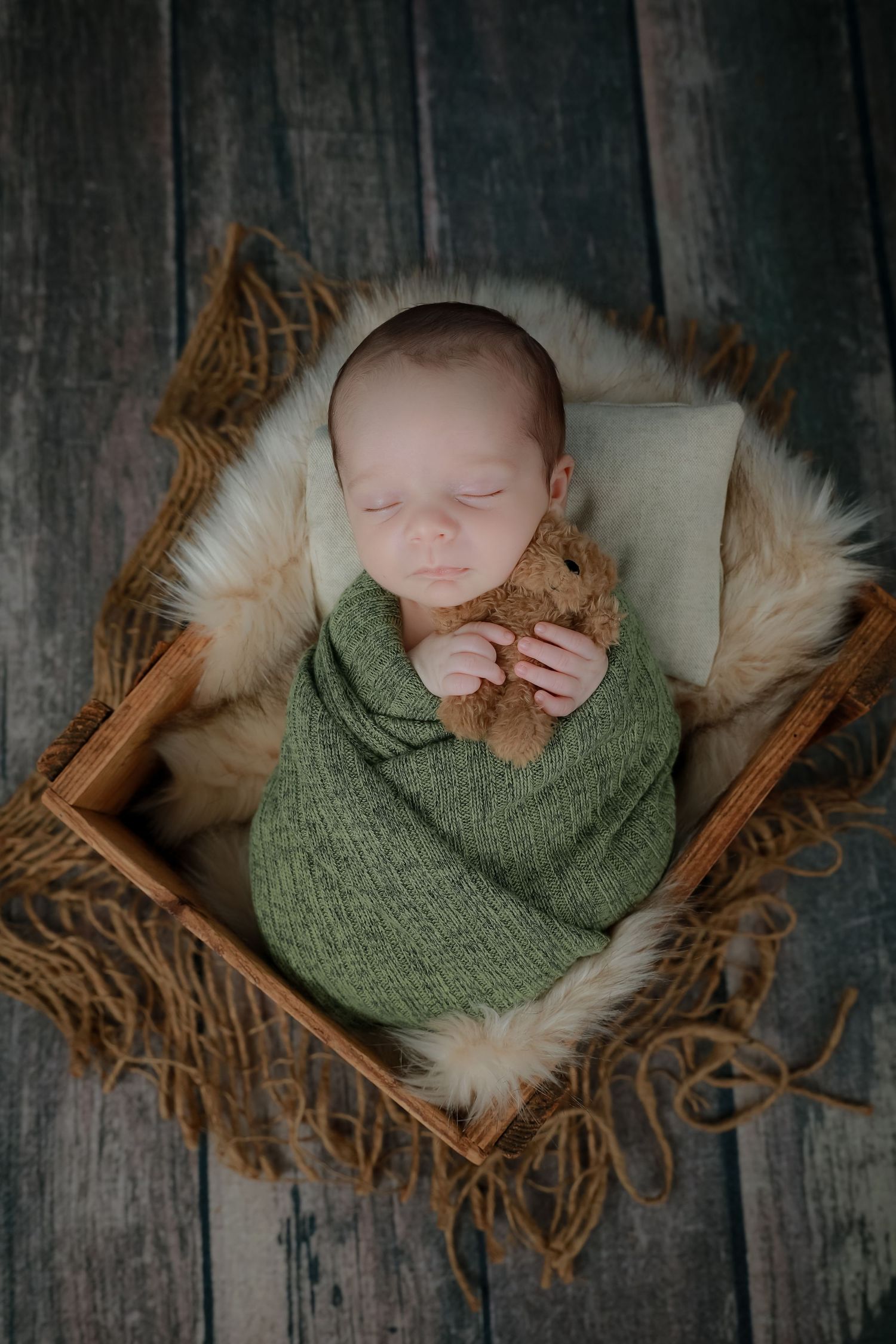 Newborn baby wrapped in green knit blanket sleeping peacefully on rustic wooden backdrop with fur accents.