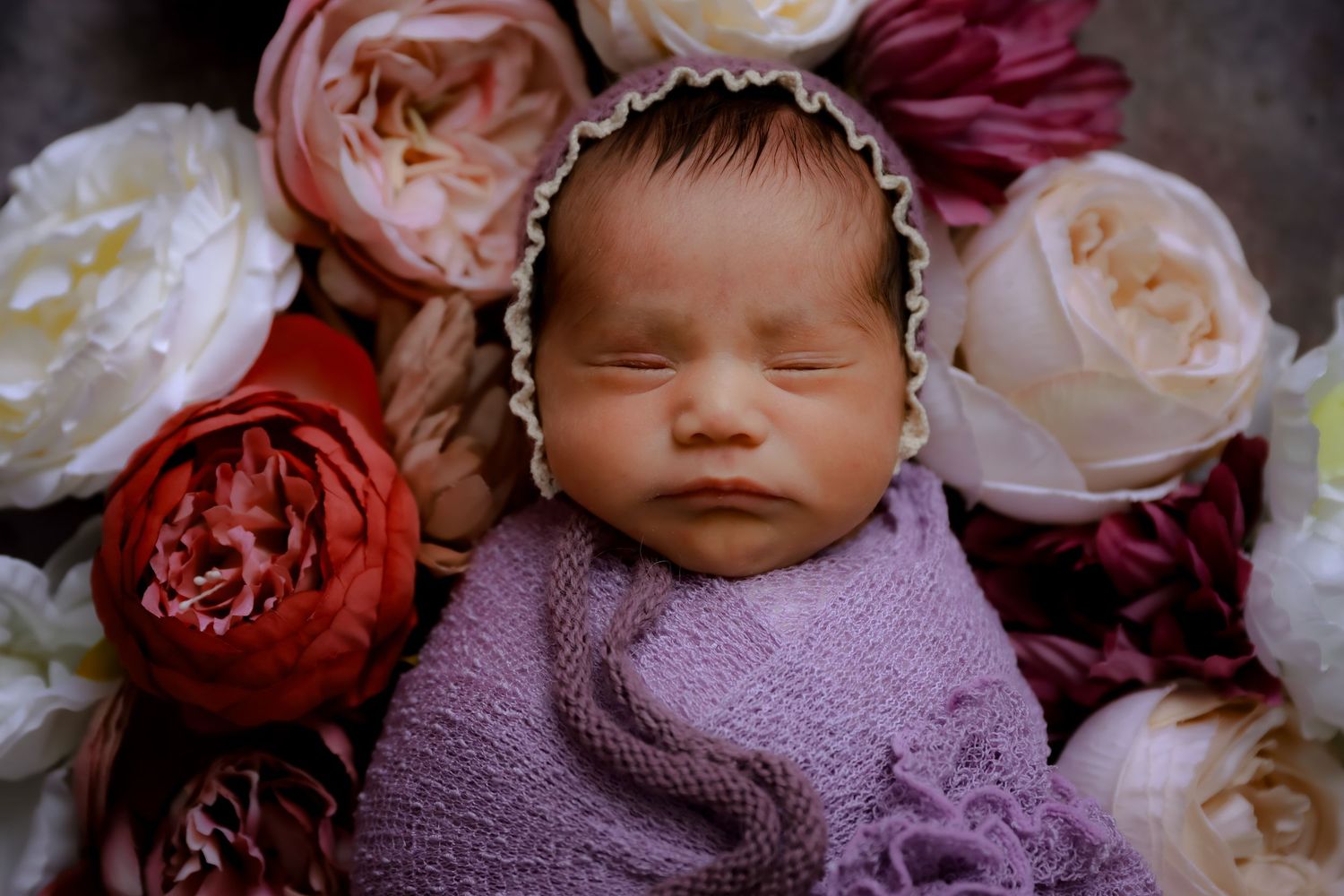 A sleeping newborn wrapped in purple knit fabric lies among pink and white roses in a professional photo setting. luxury-family-photography