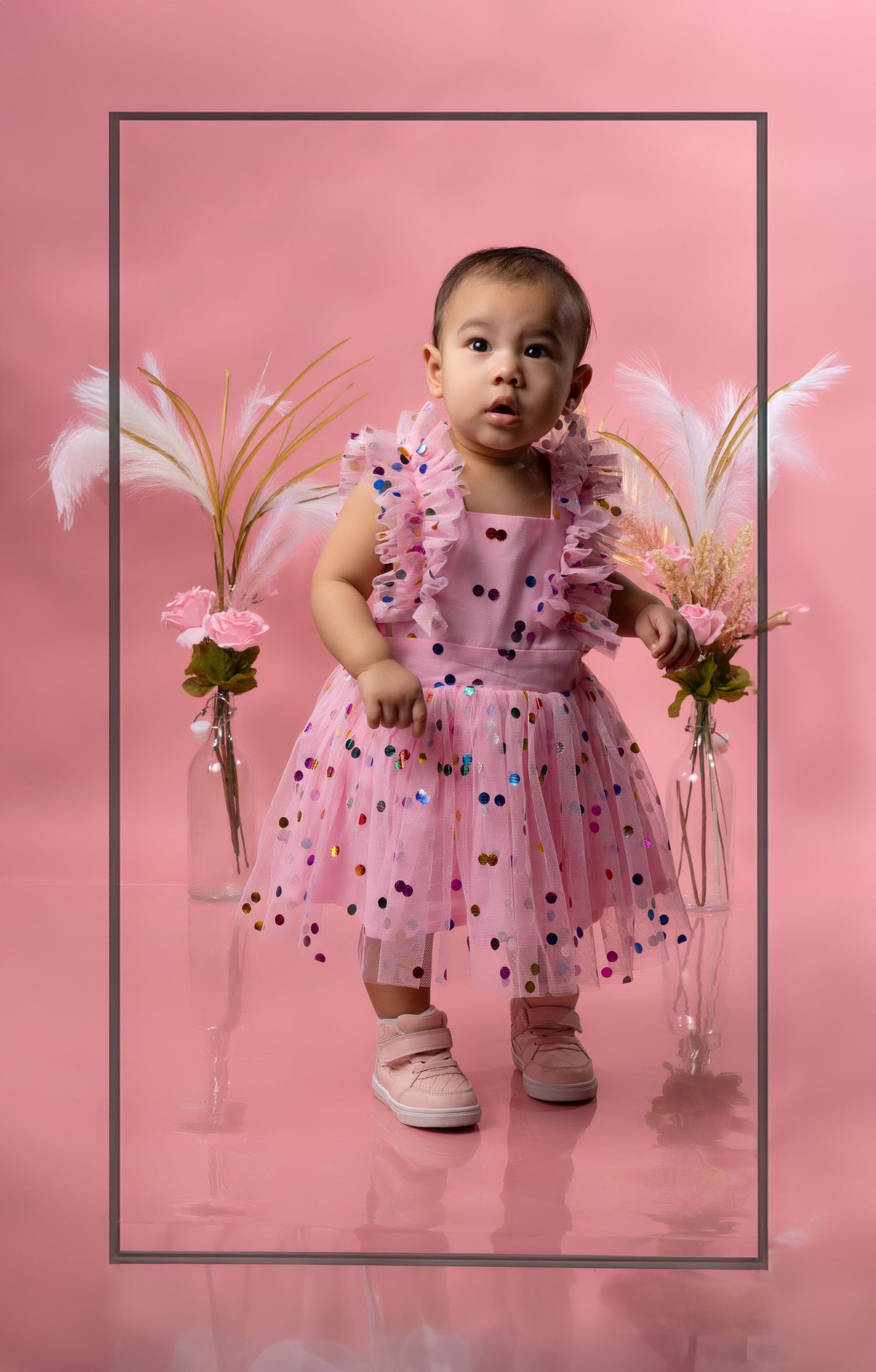 Baby wearing pink tulle dress with black polka dots poses next to floral arrangements on pink backdrop.