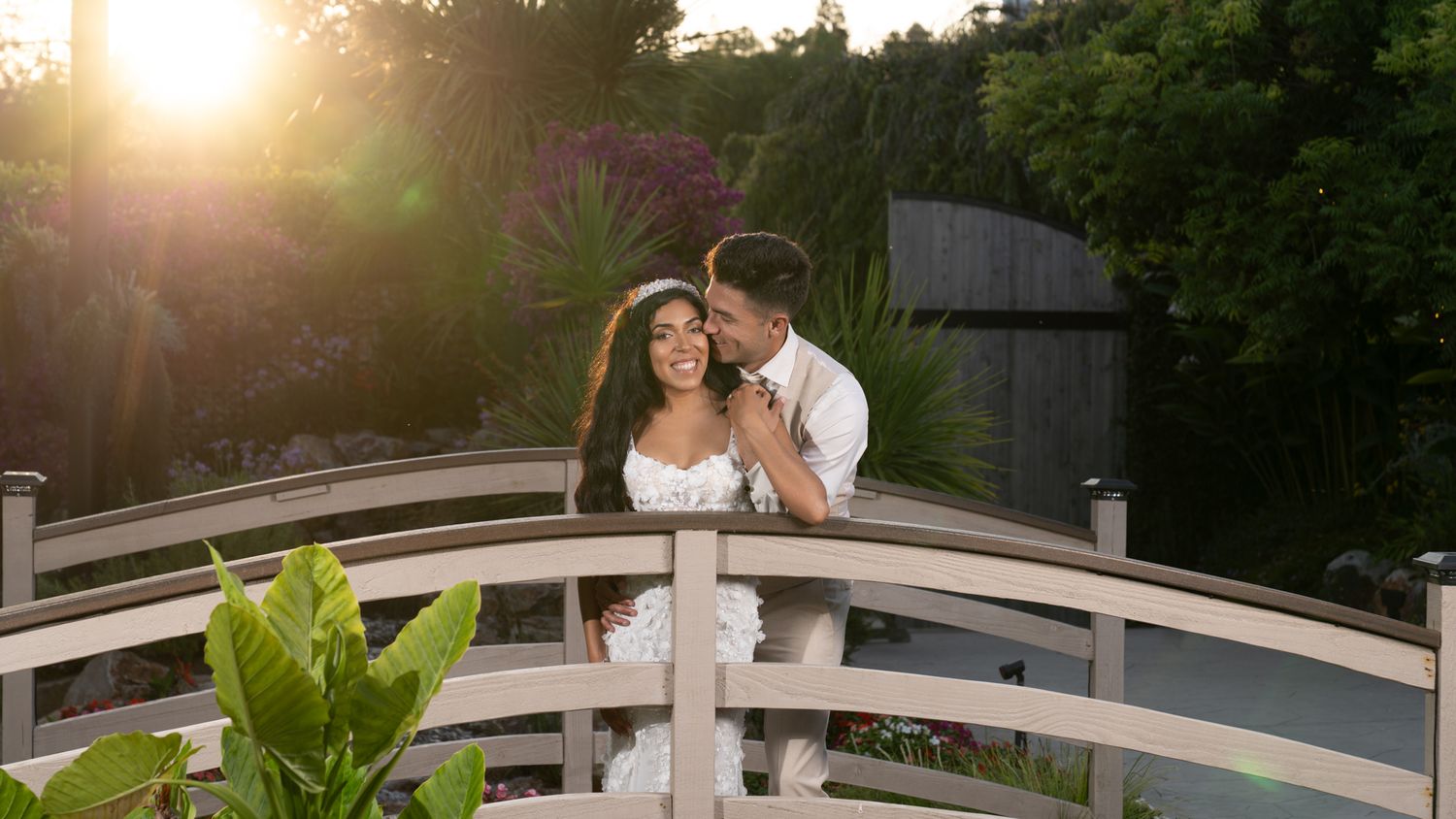 A couple embraces on a white wooden bridge during golden hour sunset in a romantic outdoor setting.