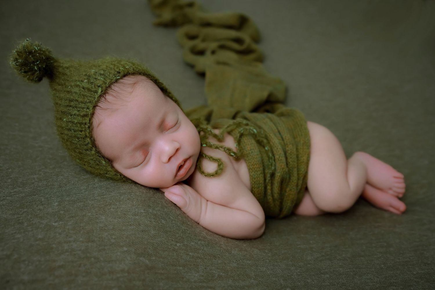 A sleeping newborn baby wearing an olive green knit outfit lies curled up on a dark textured surface. luxury-family-photography-walnut
