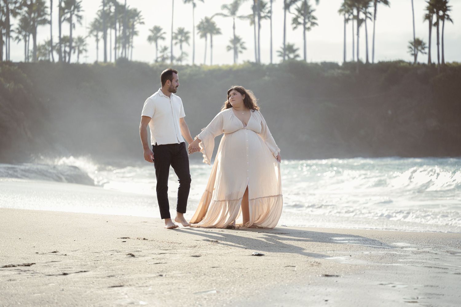 A couple sits together on a sandy beach at sunset while waves crash against the shore in the background.