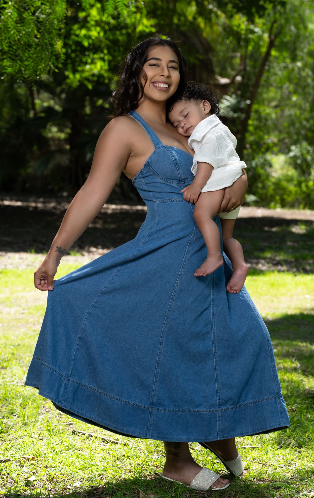 A woman in a blue denim dress twirls and dances with a small child in a white outfit on a sunny grassy lawn.