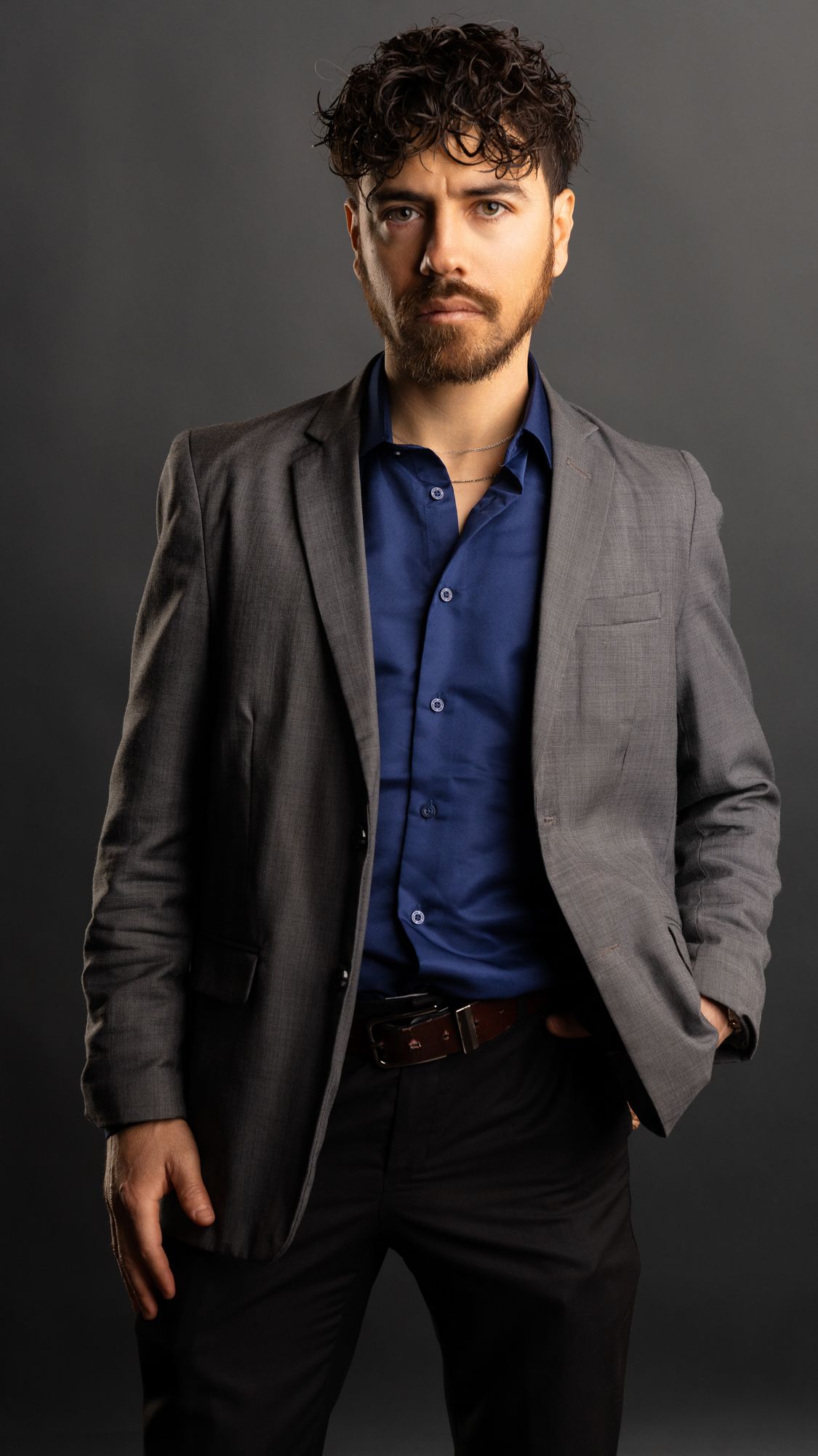 Professional studio portrait of a person wearing a gray suit jacket and navy blue dress shirt against a dark background.