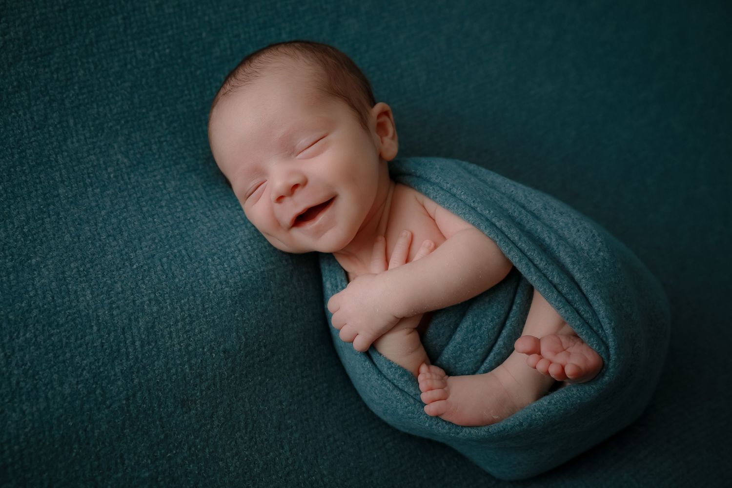 Peaceful sleeping newborn baby wrapped in soft teal blanket against dark teal background.