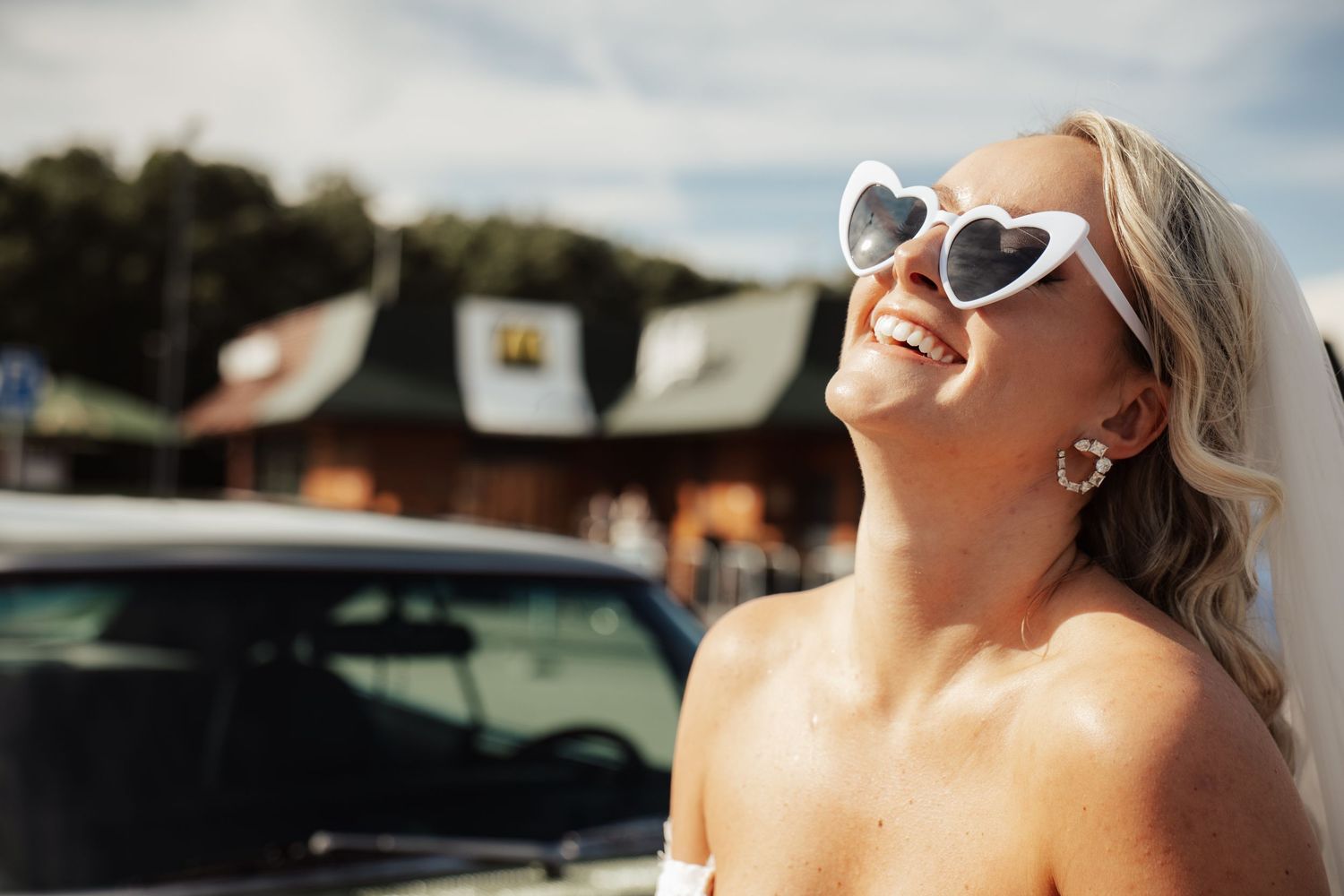 Smiling bride in white heart-shaped sunglasses laughing outdoors on a sunny day near a car.