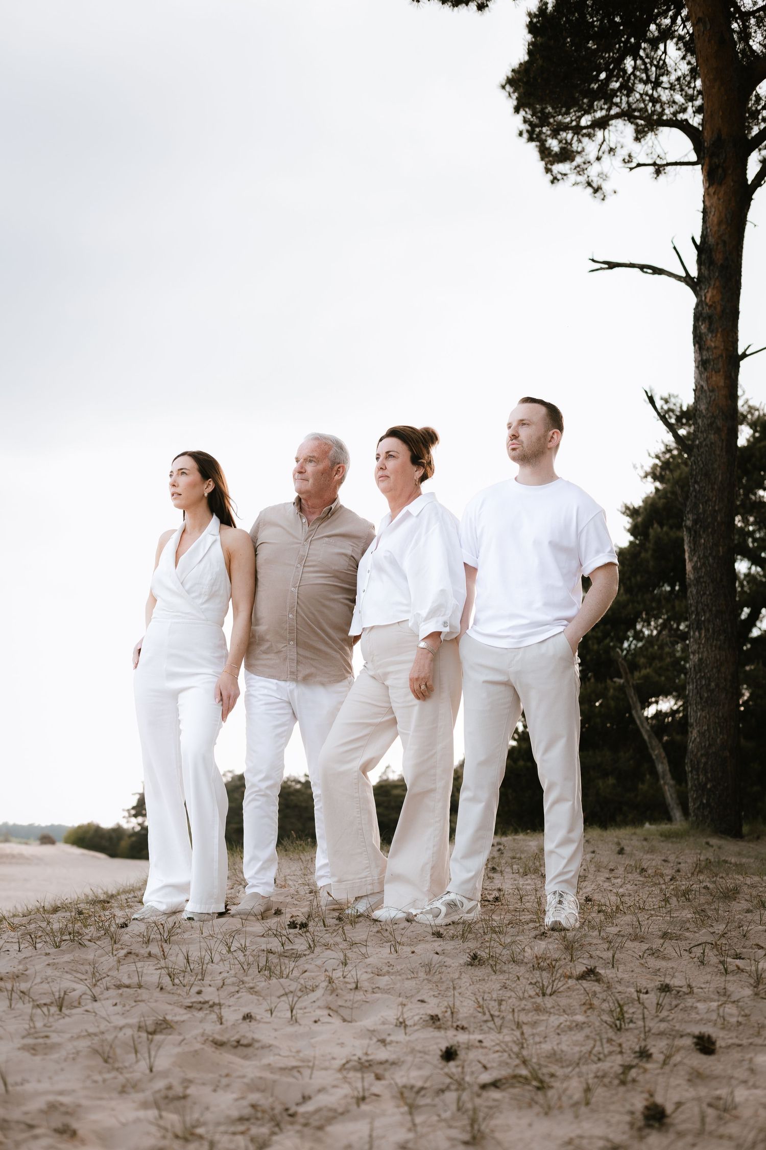 Four family members in white outfits standing on sandy ground with trees in background.