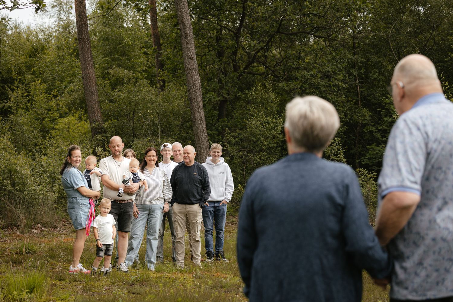 A group of people gathered outdoors in a forest clearing, with two figures in the foreground facing the crowd.
