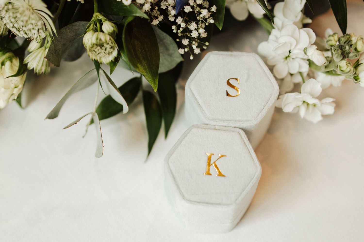 White hexagonal ring boxes with gold initials S and K surrounded by delicate white flowers.