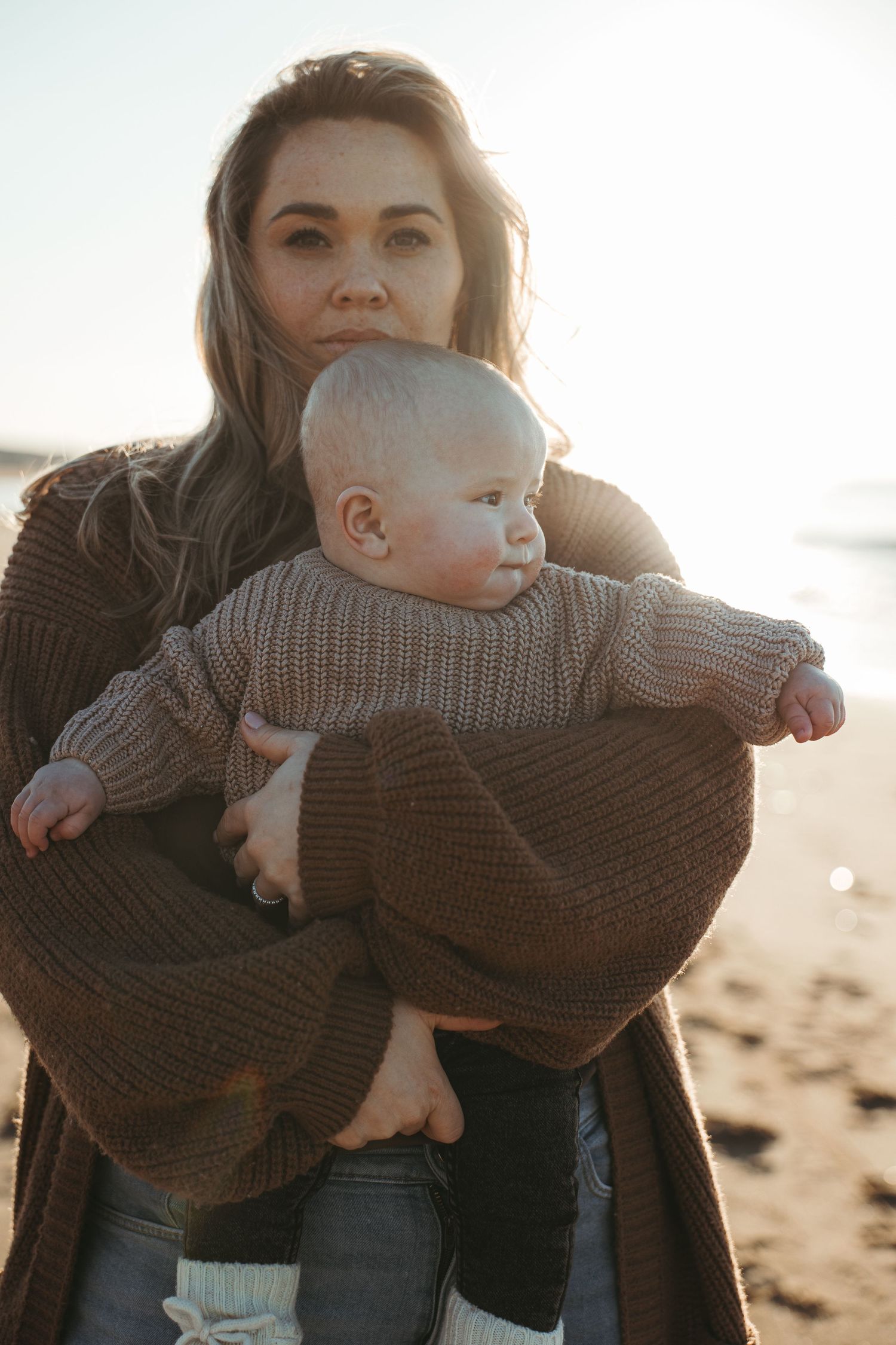 A parent holds a baby wearing a knit sweater on a windswept beach at sunset.