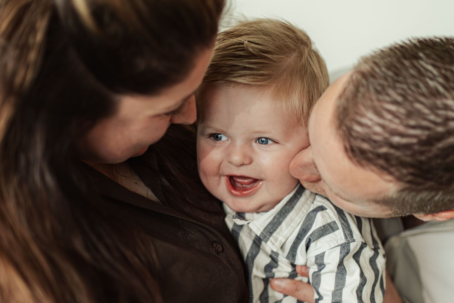 Happy baby boy smiling while parents kiss his cheeks in a warm family portrait moment.