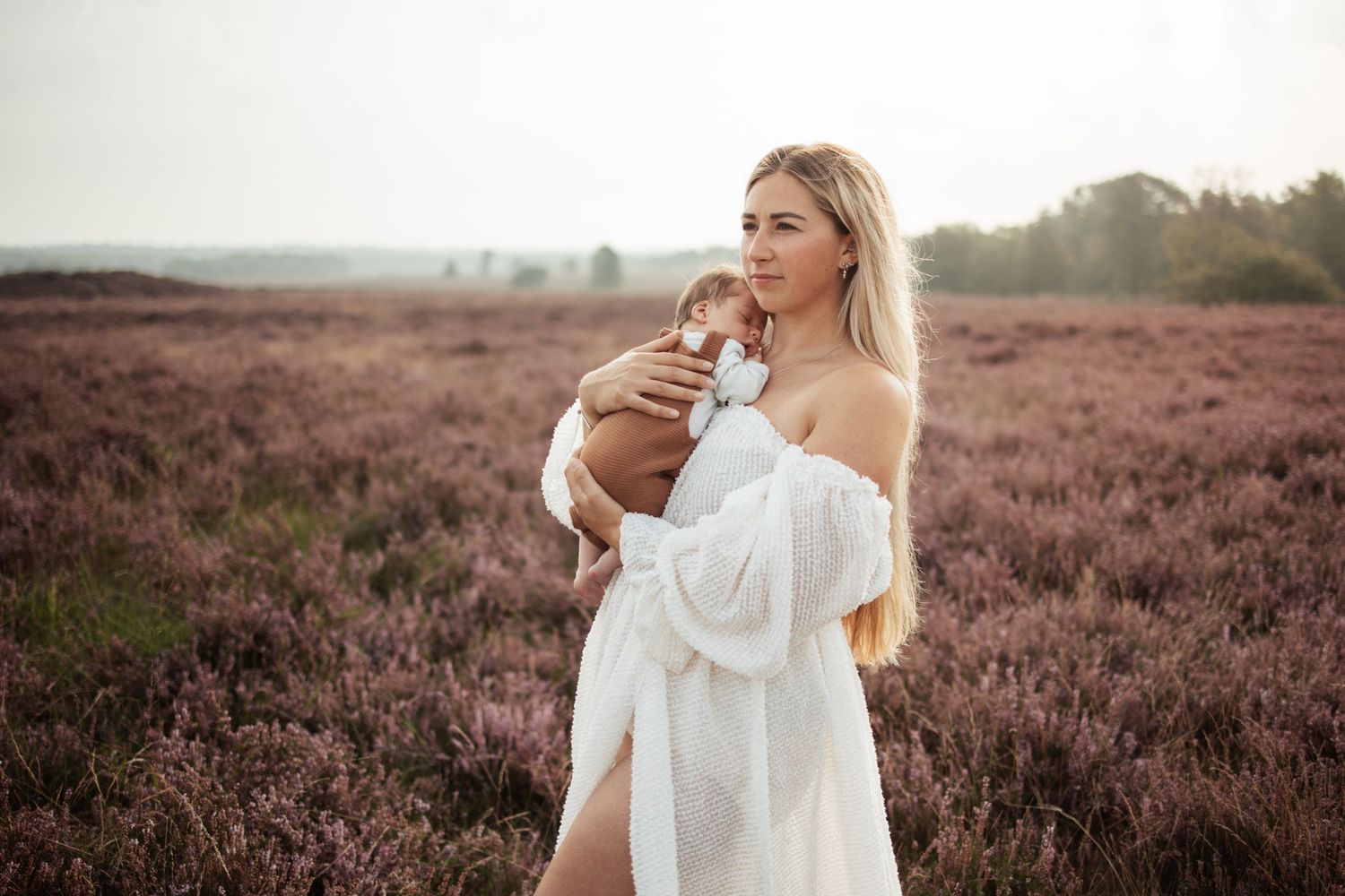 Woman in flowing white dress standing in field of purple heather at sunset.