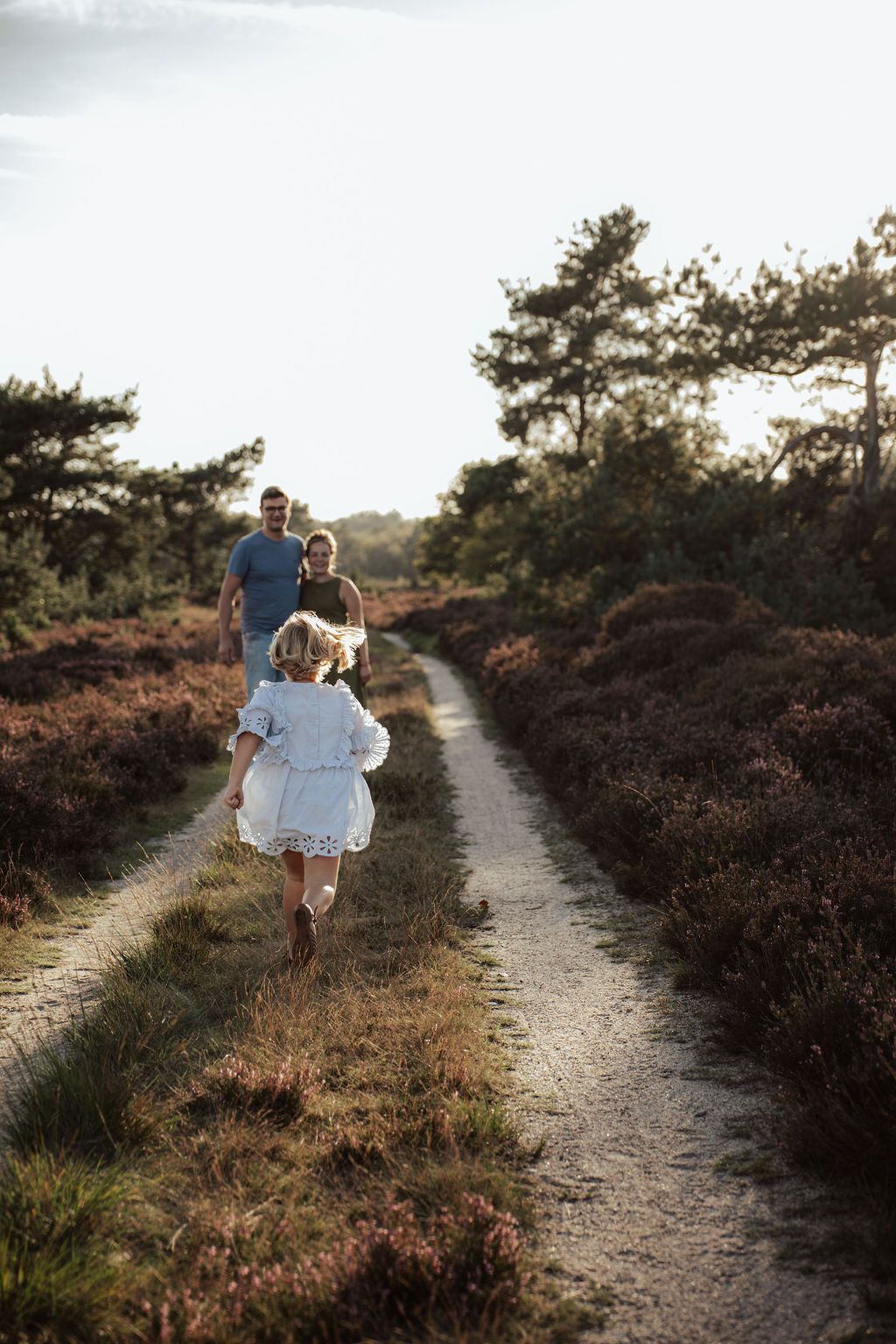 People walking along a scenic dirt trail surrounded by blooming heather at sunset.