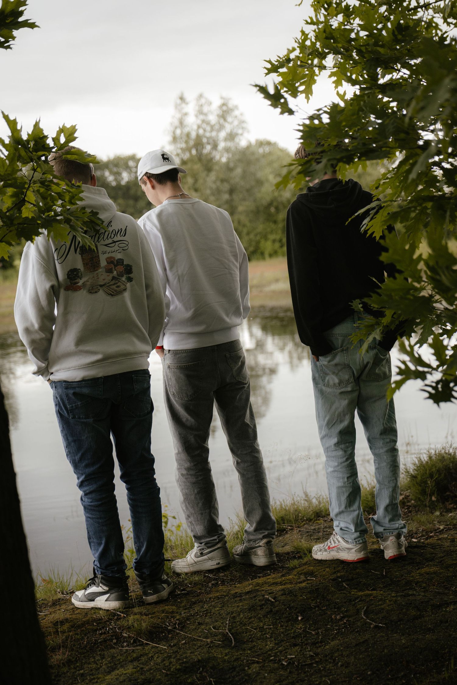 Three young men standing with their backs turned, overlooking a calm lake surrounded by lush green trees.