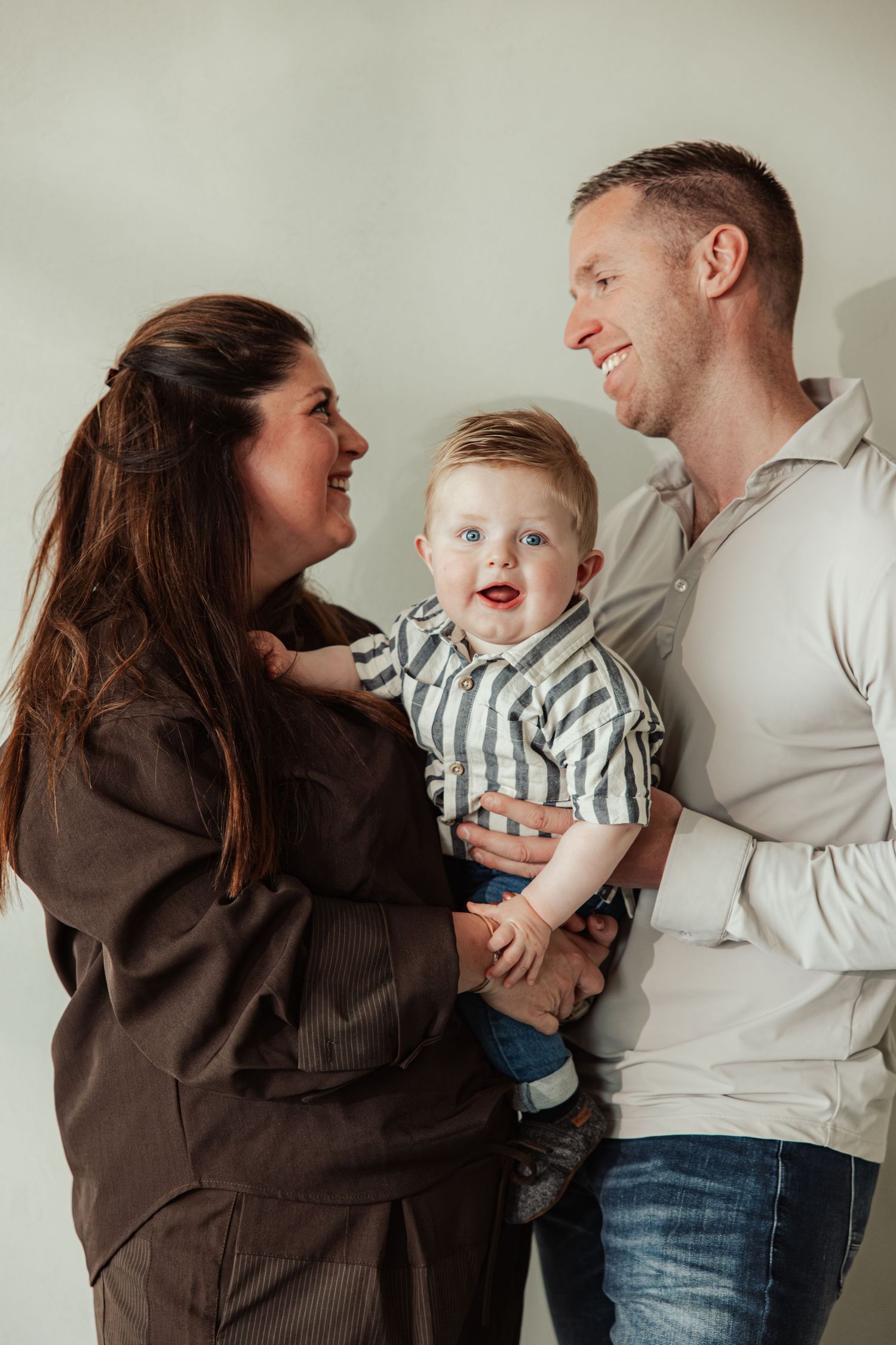 Happy family portrait with mother, father, and smiling baby boy in striped shirt against light background.