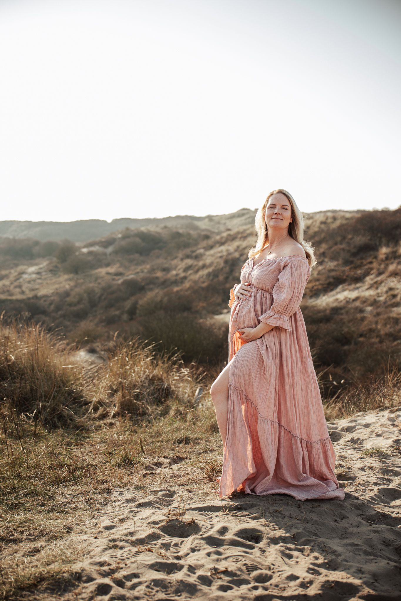 Maternity portrait in a flowing pink dress against sandy dunes at golden hour.