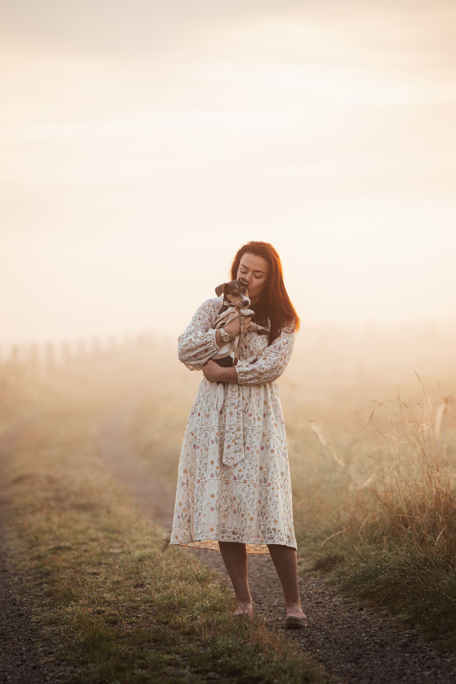 A person in a white floral dress stands in a misty field during golden hour creating a dreamy atmosphere.