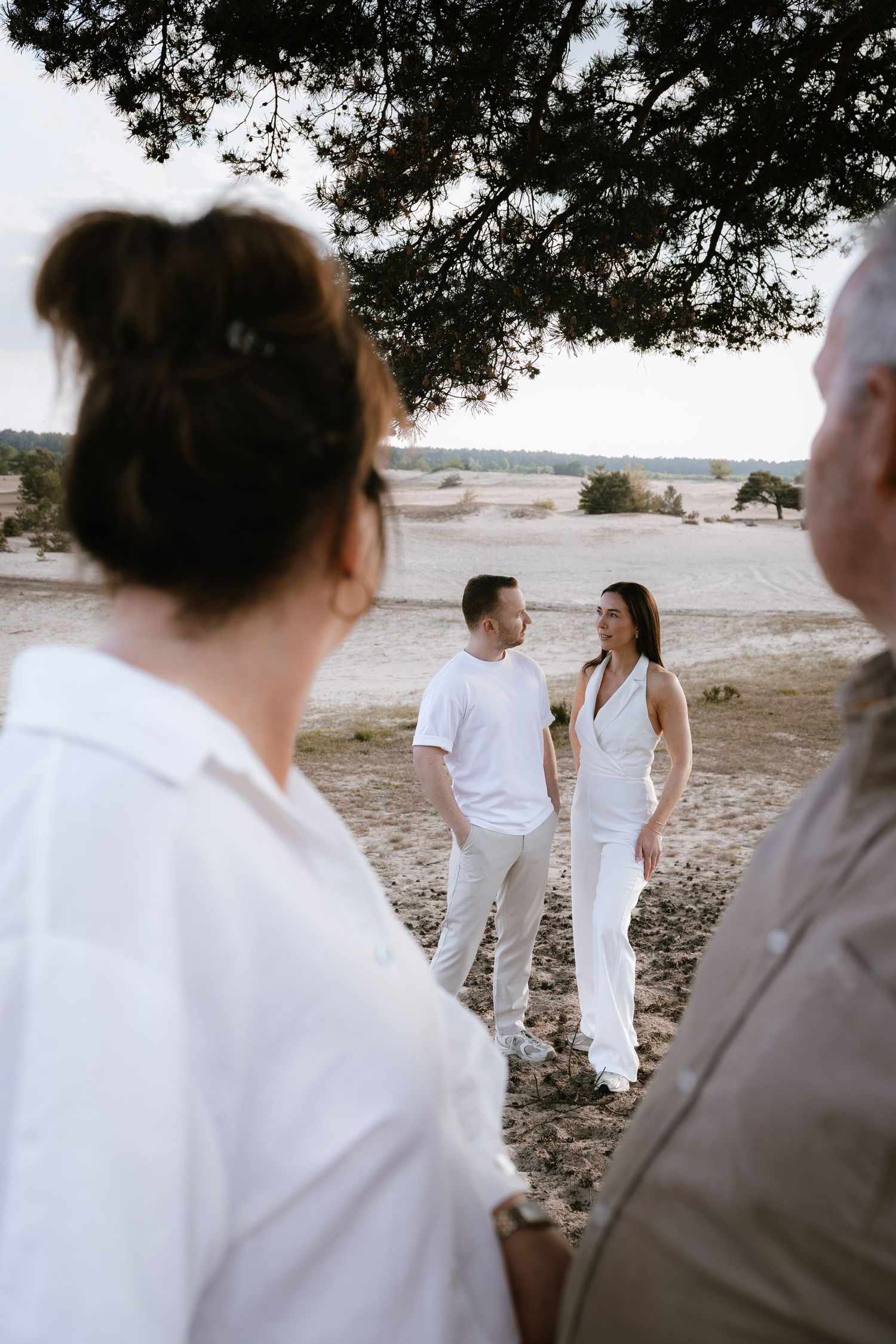 Couple in white outfits standing together in sandy landscape, observed by guests during an outdoor ceremony.
