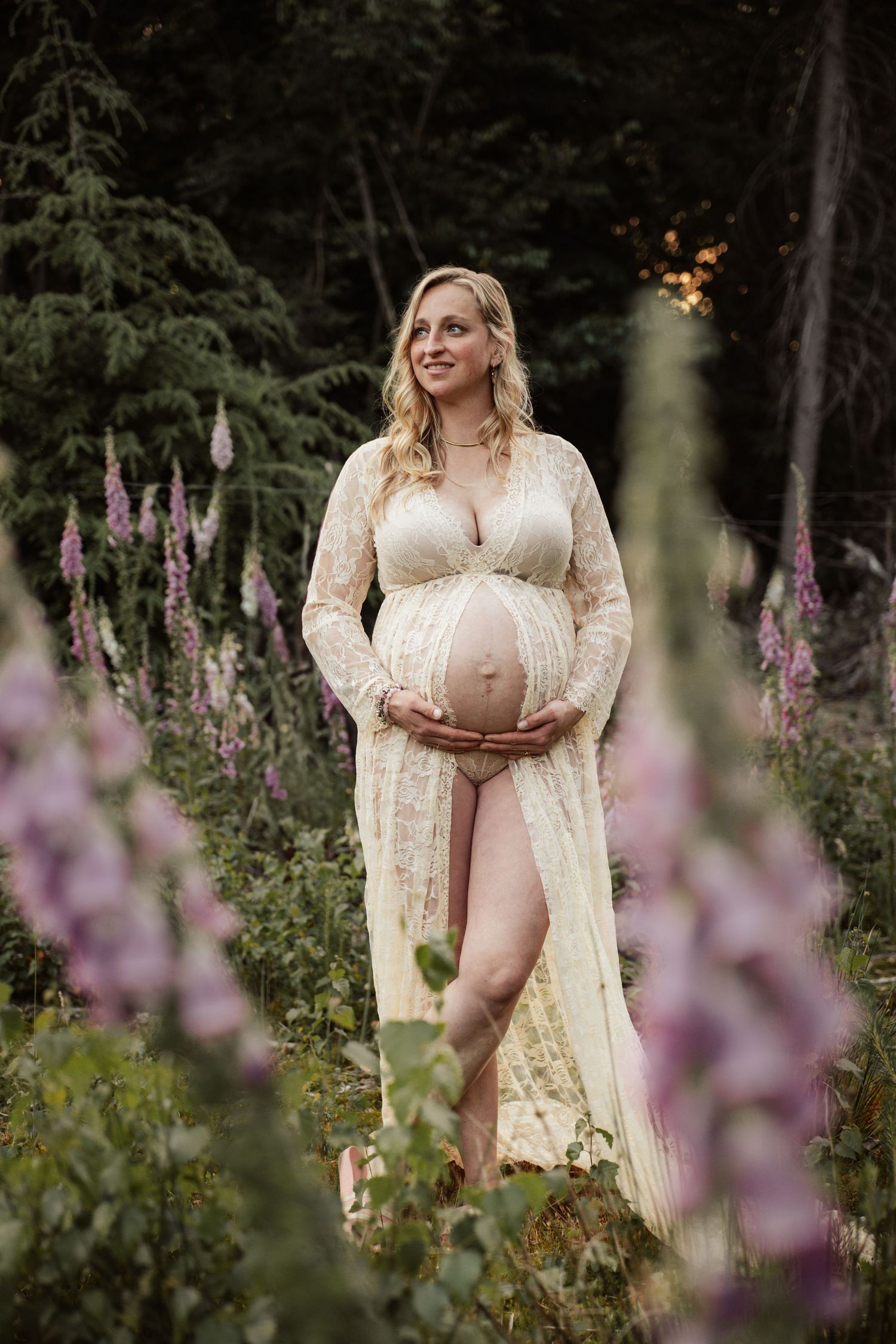 Maternity portrait in lace robe surrounded by purple foxglove flowers in garden setting.