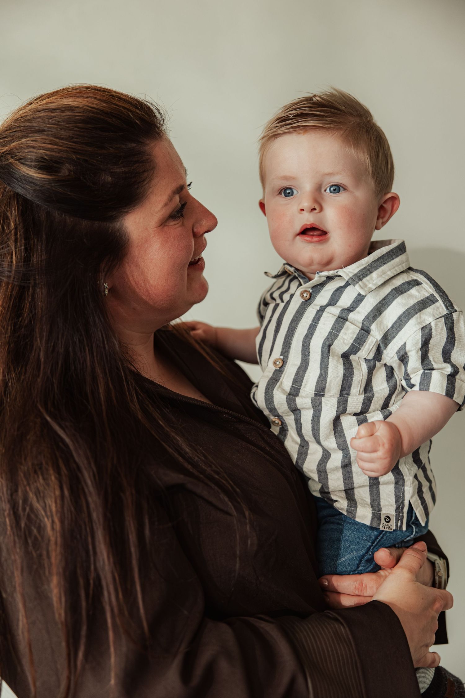 A woman with long dark hair holds a blue-eyed baby boy in a striped shirt, smiling warmly at him.