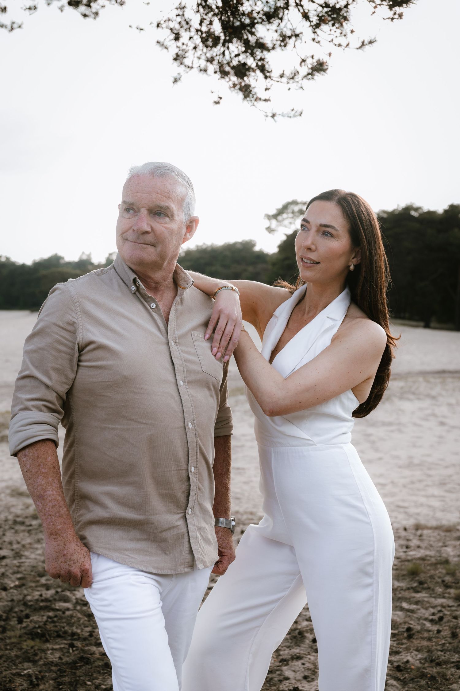 An older man and younger woman pose together outdoors on a sandy beach, wearing neutral and white outfits.