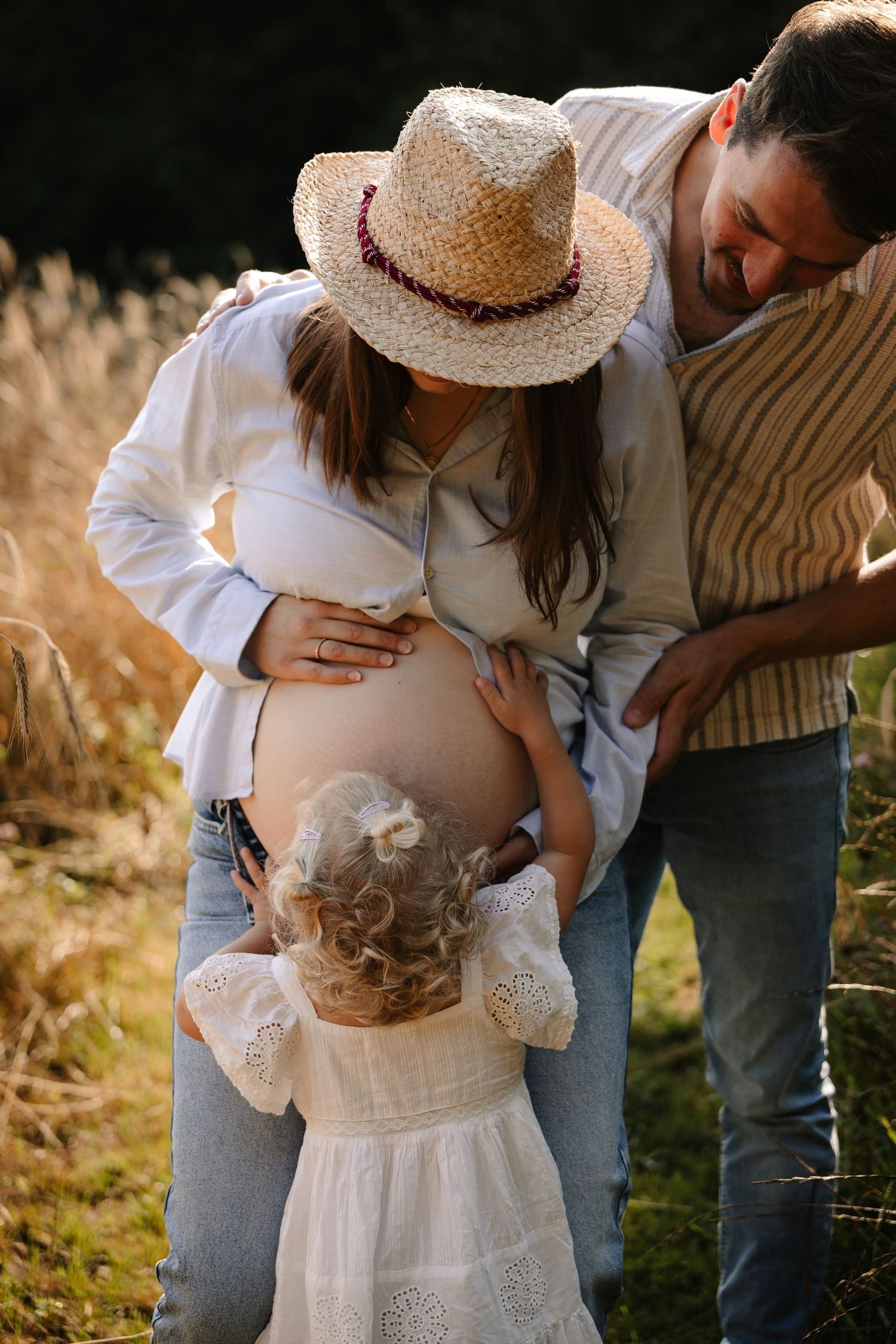Pregnant woman in straw hat with husband and toddler daughter touching baby bump outdoors in golden sunlight.