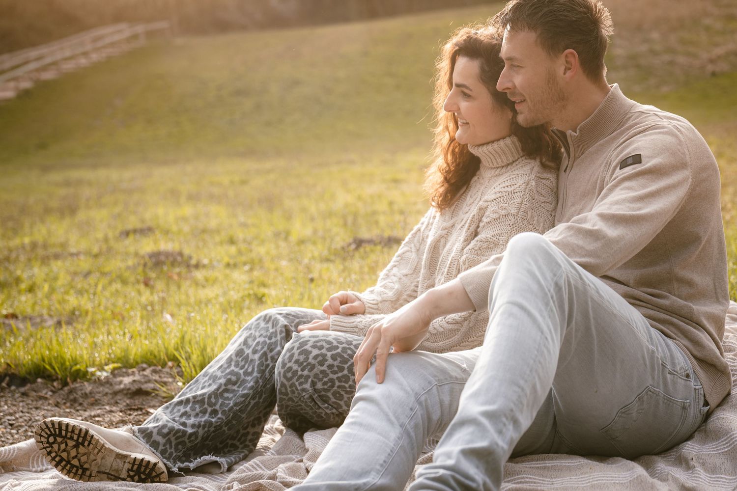Couple sitting together on a blanket in a sunny park, sharing a romantic moment outdoors in casual sweaters.