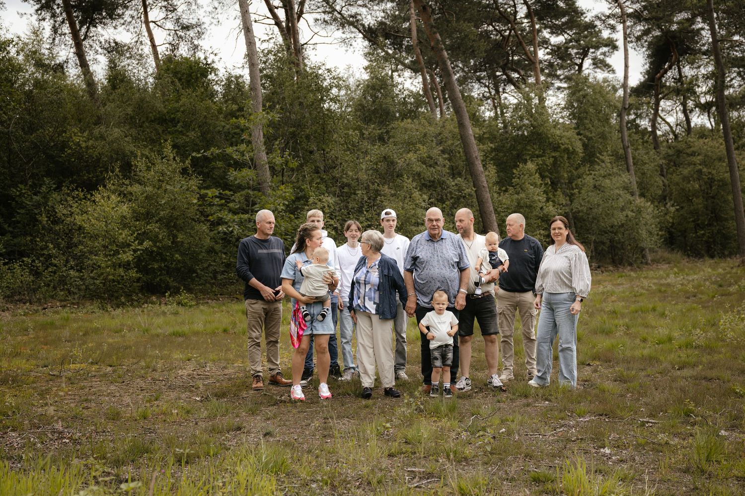 Multi-generational family group portrait outdoors in a forest clearing surrounded by tall pine trees and lush greenery.