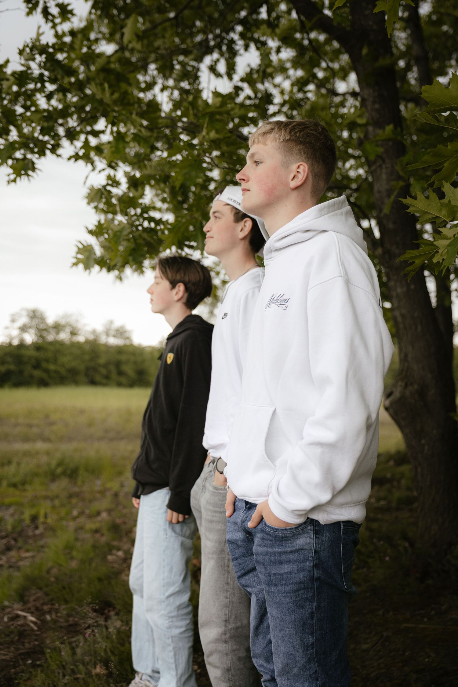 Three teenage boys in hoodies standing outdoors near a tree, gazing into the distance on a cloudy day.