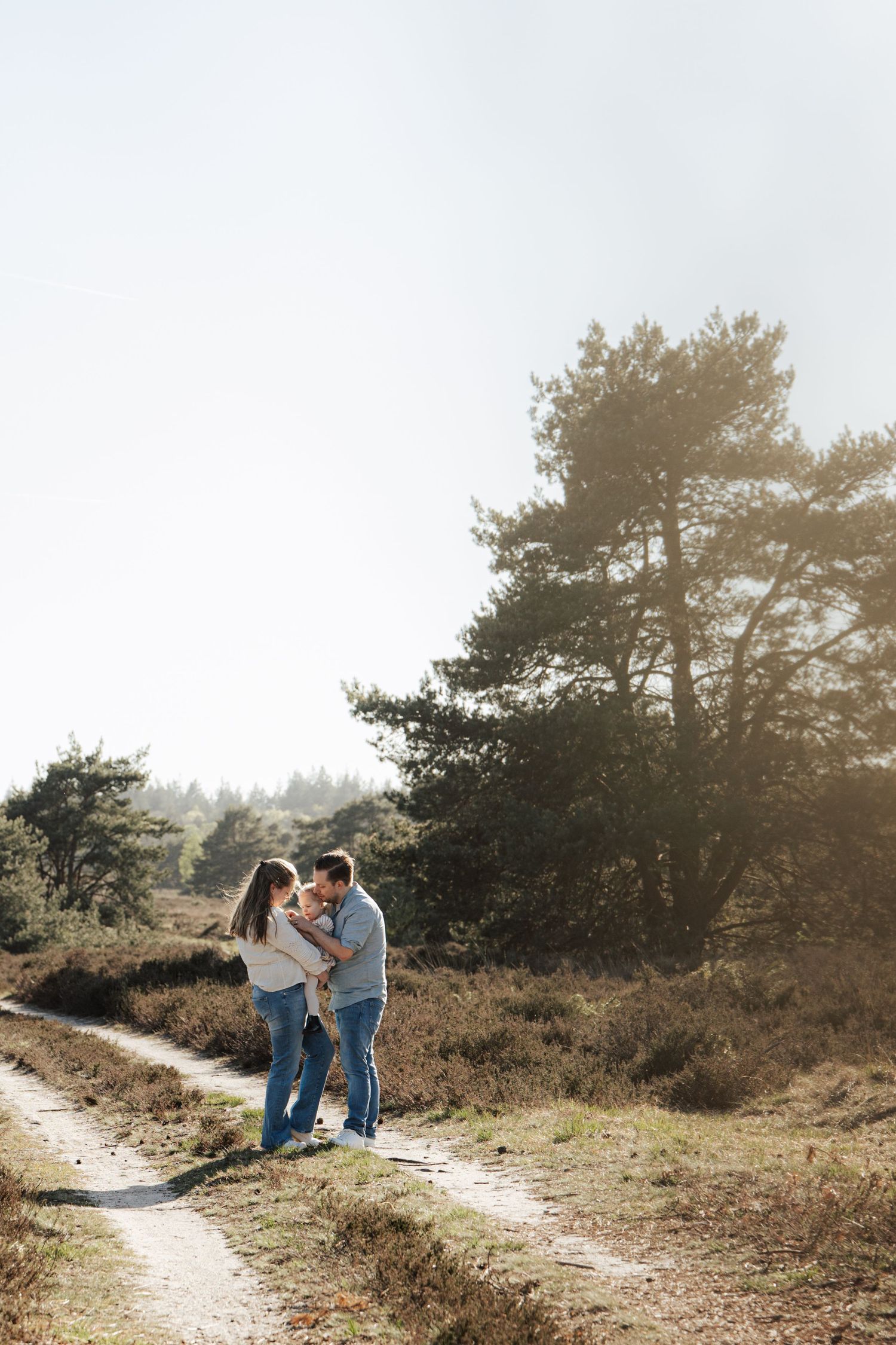 A young family with a baby stands on a sunny dirt path surrounded by heathland and pine trees.