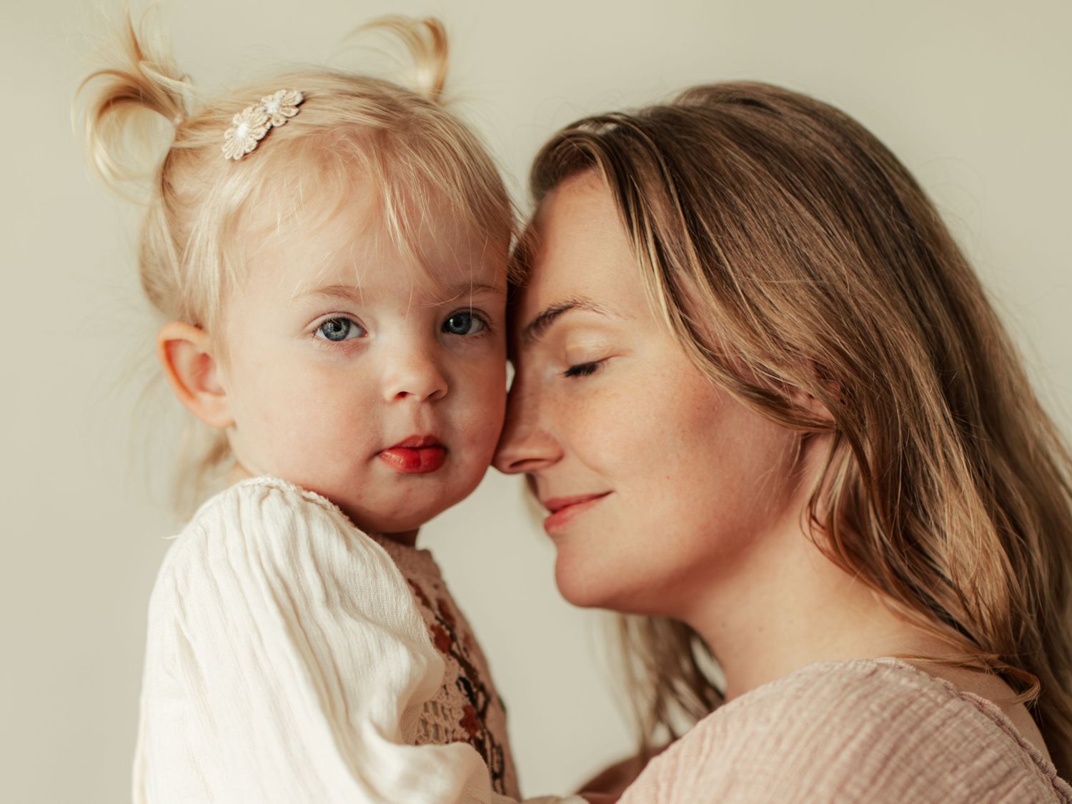 A tender moment between a mother and her blonde toddler daughter, gently touching foreheads in a warm embrace.