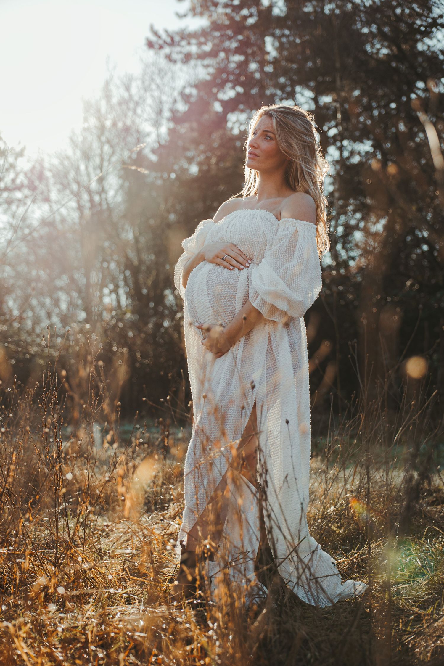 Ethereal maternity portrait of woman in white dress among tall grass at sunset.