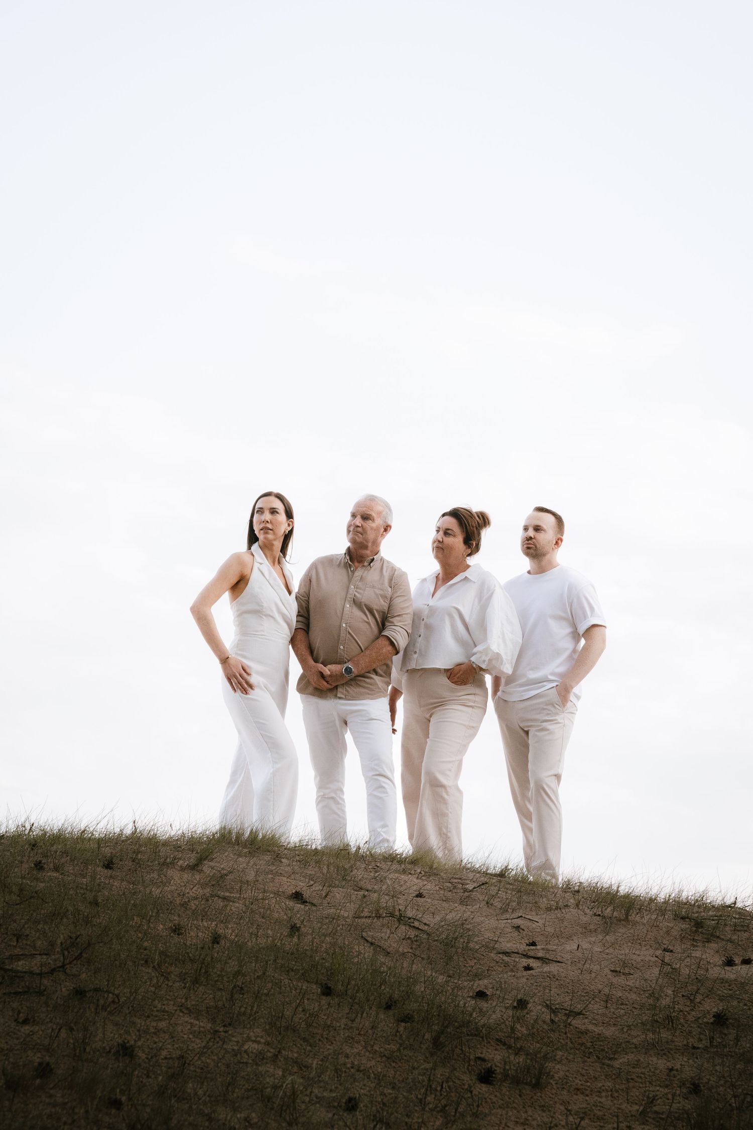 Family of four dressed in white standing on grassy hill against overcast sky.